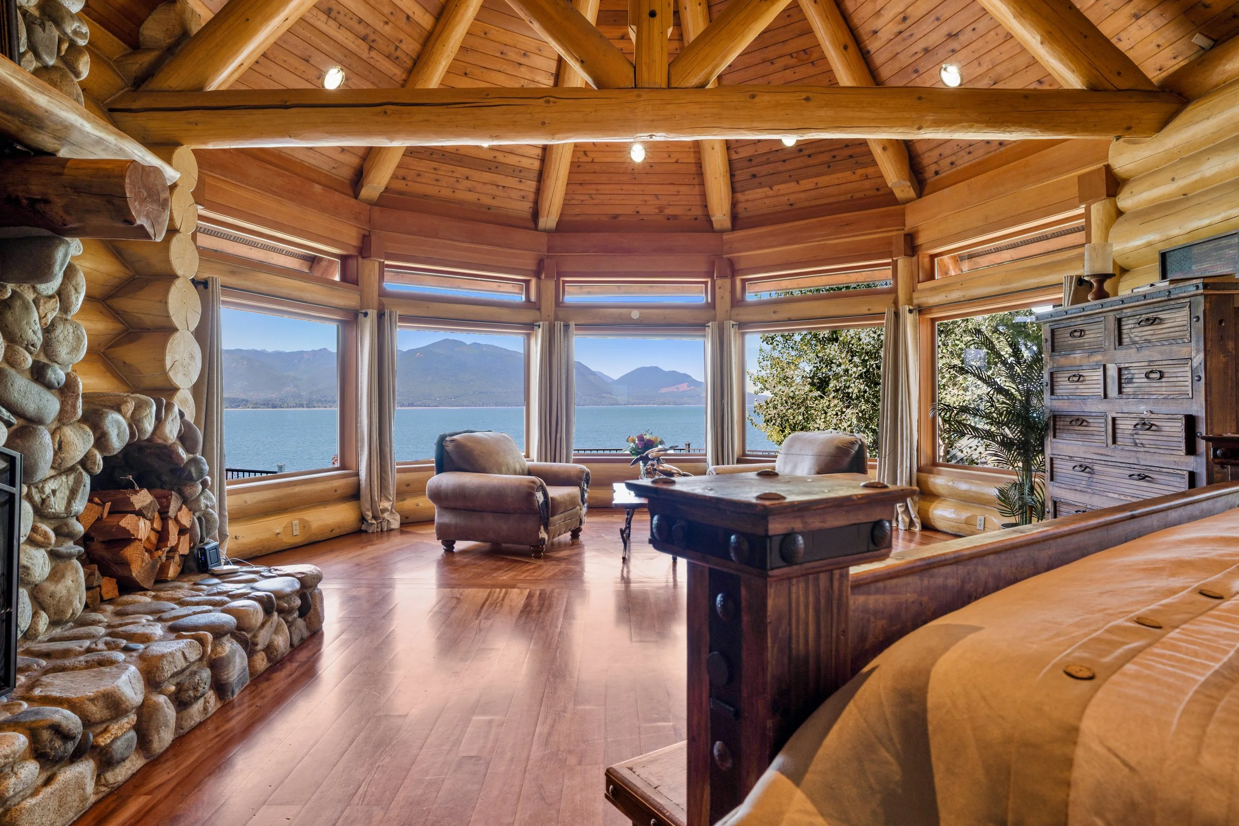 Cozy log cabin living room with large windows overlooking a mountain lake, featuring wooden walls and ceiling, a stone fireplace, and rustic furniture.