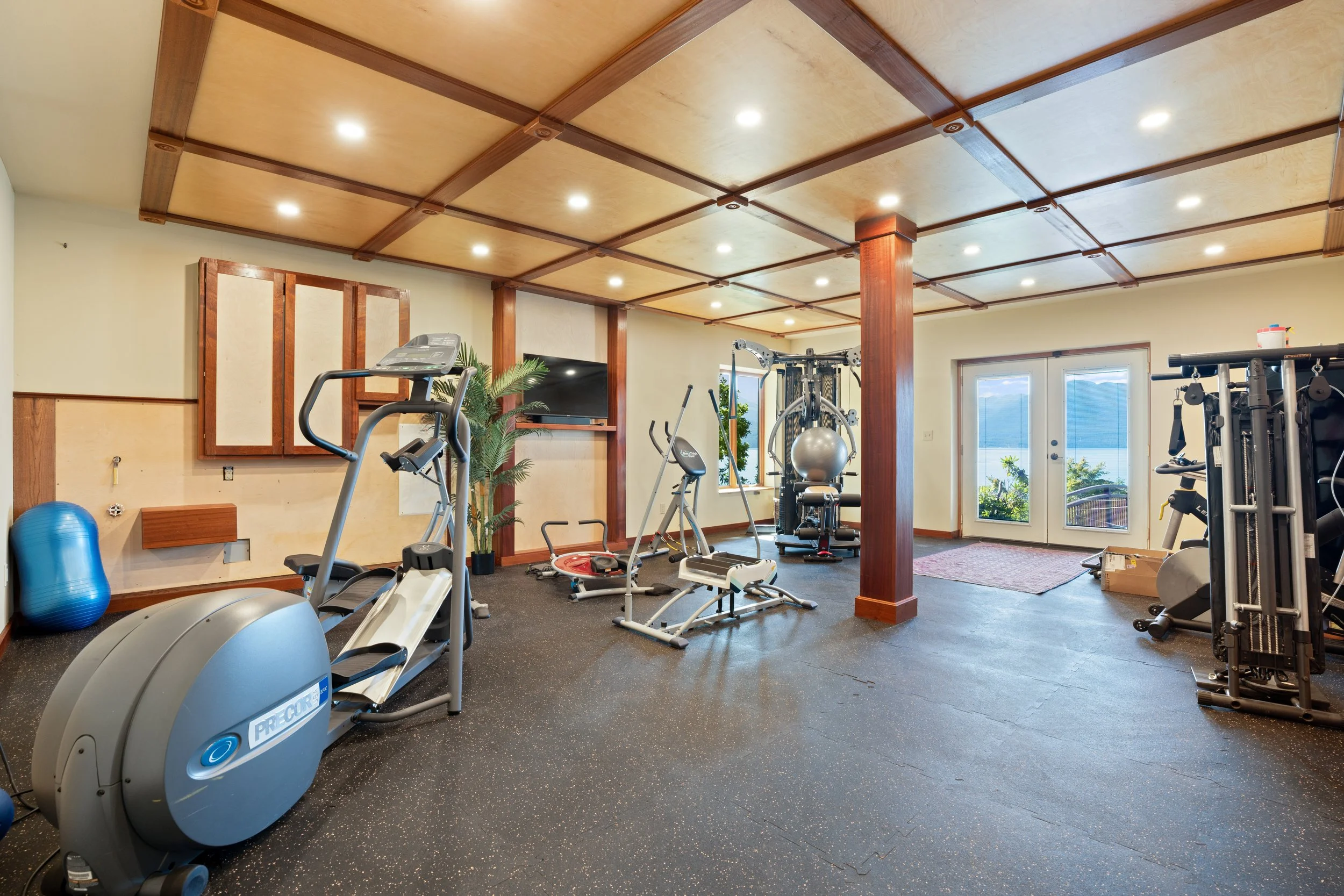 Home gym with various exercise equipment including a treadmill, elliptical, stationary bike, weights, and a stability ball, with wooden ceiling beams and large windows showing an outdoor view.