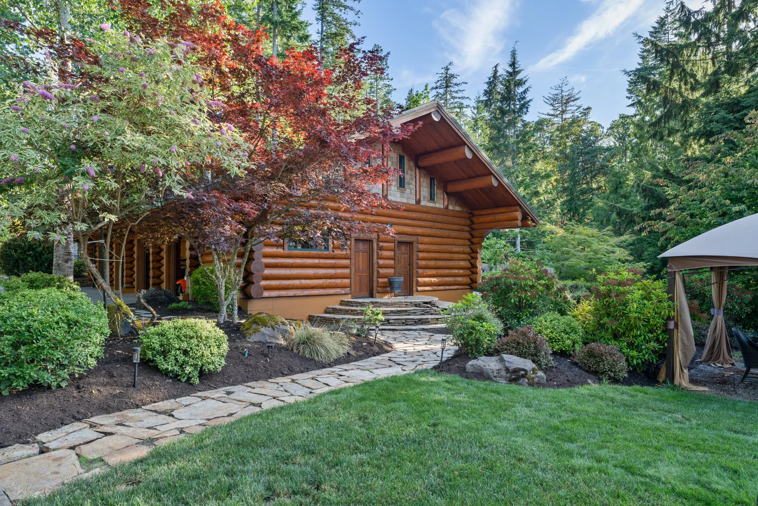 A wooden log cabin surrounded by a lush garden with a stone pathway, green grass, trees, and a canopy with curtains on the right.