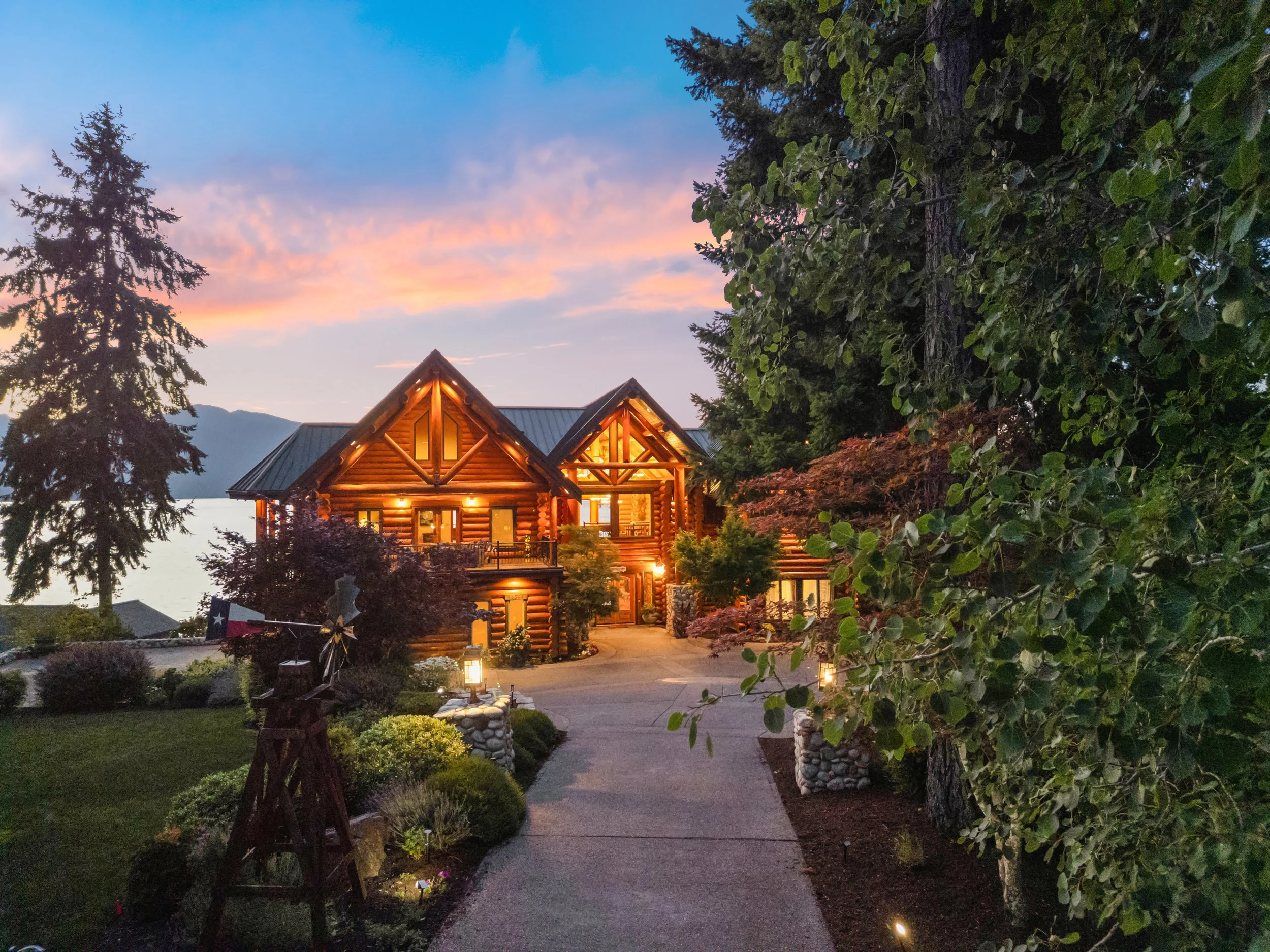 A cozy log cabin house illuminated at night in a wooded area, with a paved pathway, landscaped garden, trees, and a scenic lake with mountains in the background, under a colorful sunset sky.