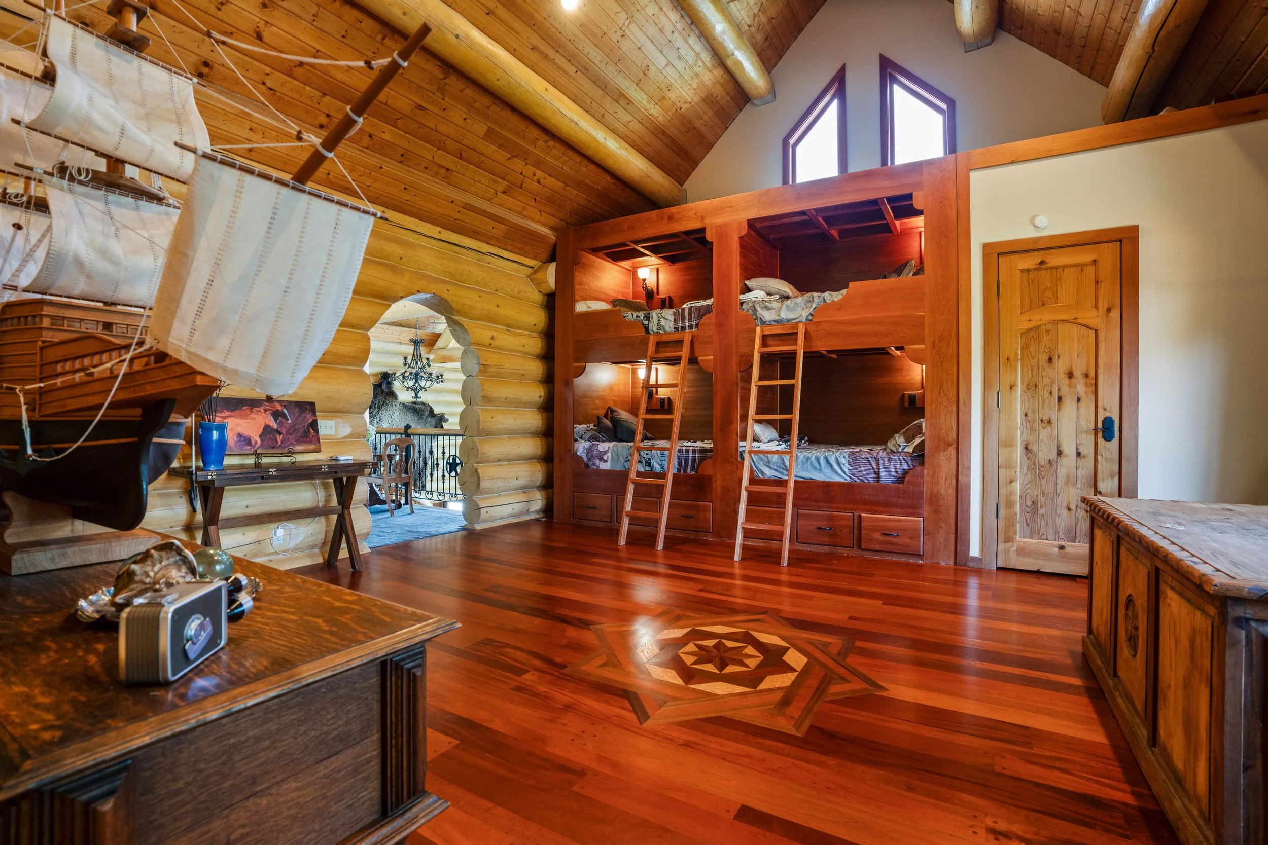 Interior of a log cabin with a wooden ceiling, two windows, and a bunk bed with four mattresses. There is a wooden door, a table with books and decorative items, and a mounted animal head on the wall.