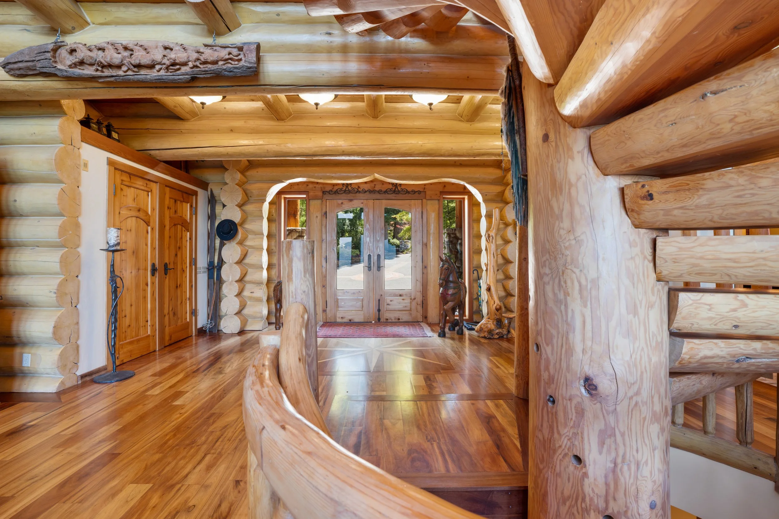 Interior view of a log cabin entry hall with wooden walls, ceiling and floors, a glass double door leading outside, and decorative items including a carved wooden horse figure and a dark hat on a stand.