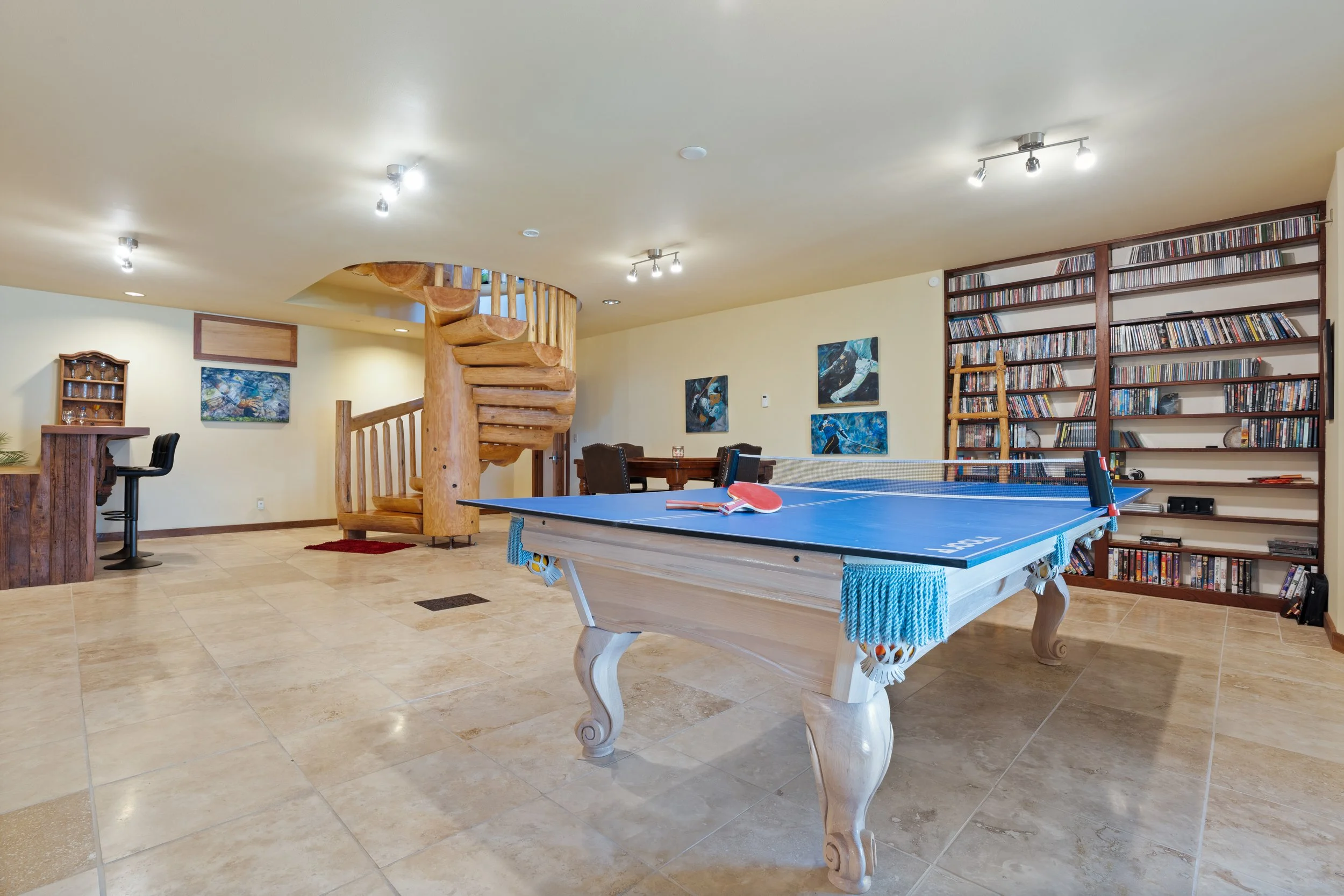 A recreational room with a wooden spiral staircase, a blue ping pong table with paddles and a net, a large bookshelf filled with DVDs or CDs, a bar area with barstools, and colorful artwork on the walls.