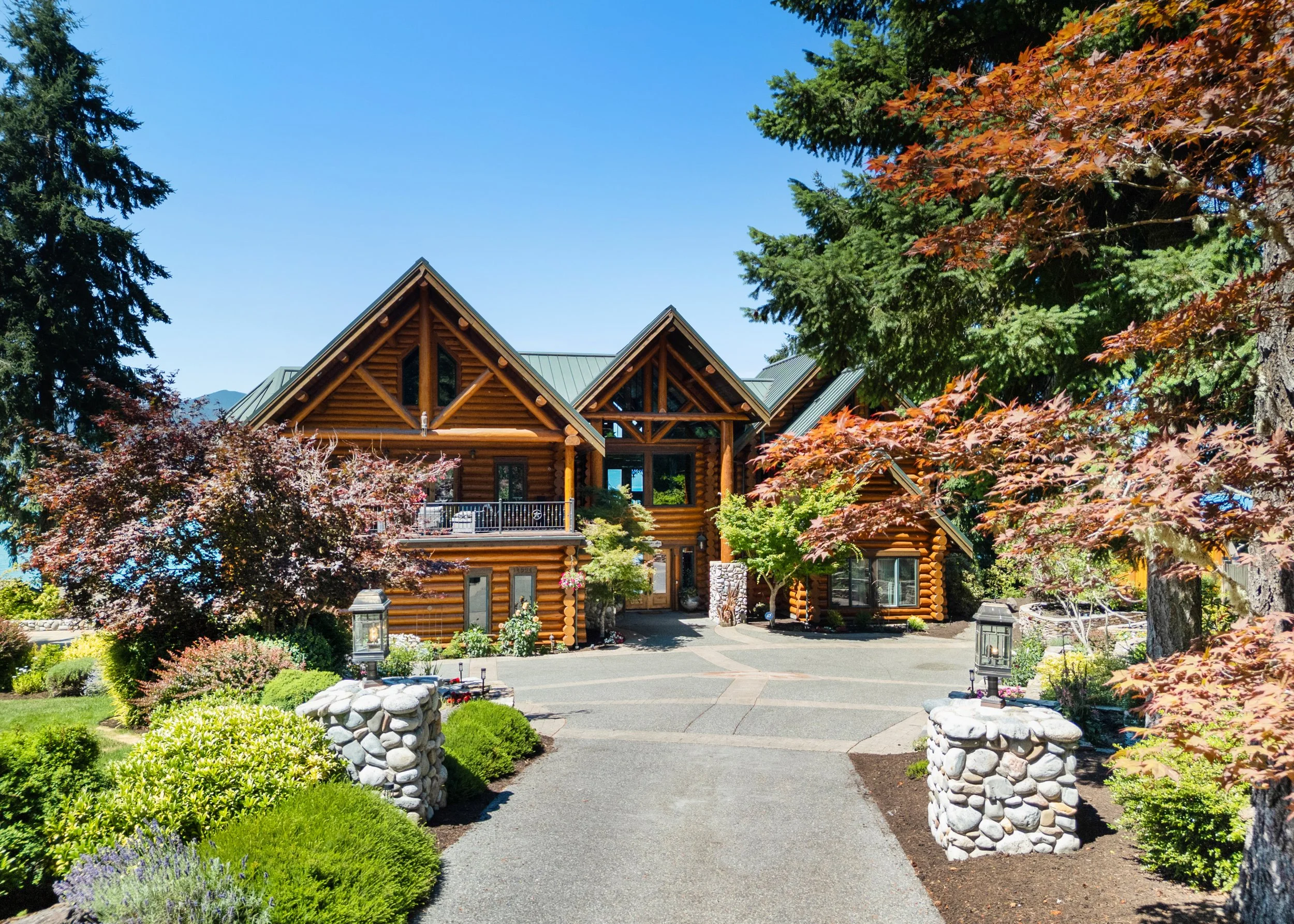 A log-style house with large windows, surrounded by a well-maintained garden with colorful plants and trees, and a clear blue sky.