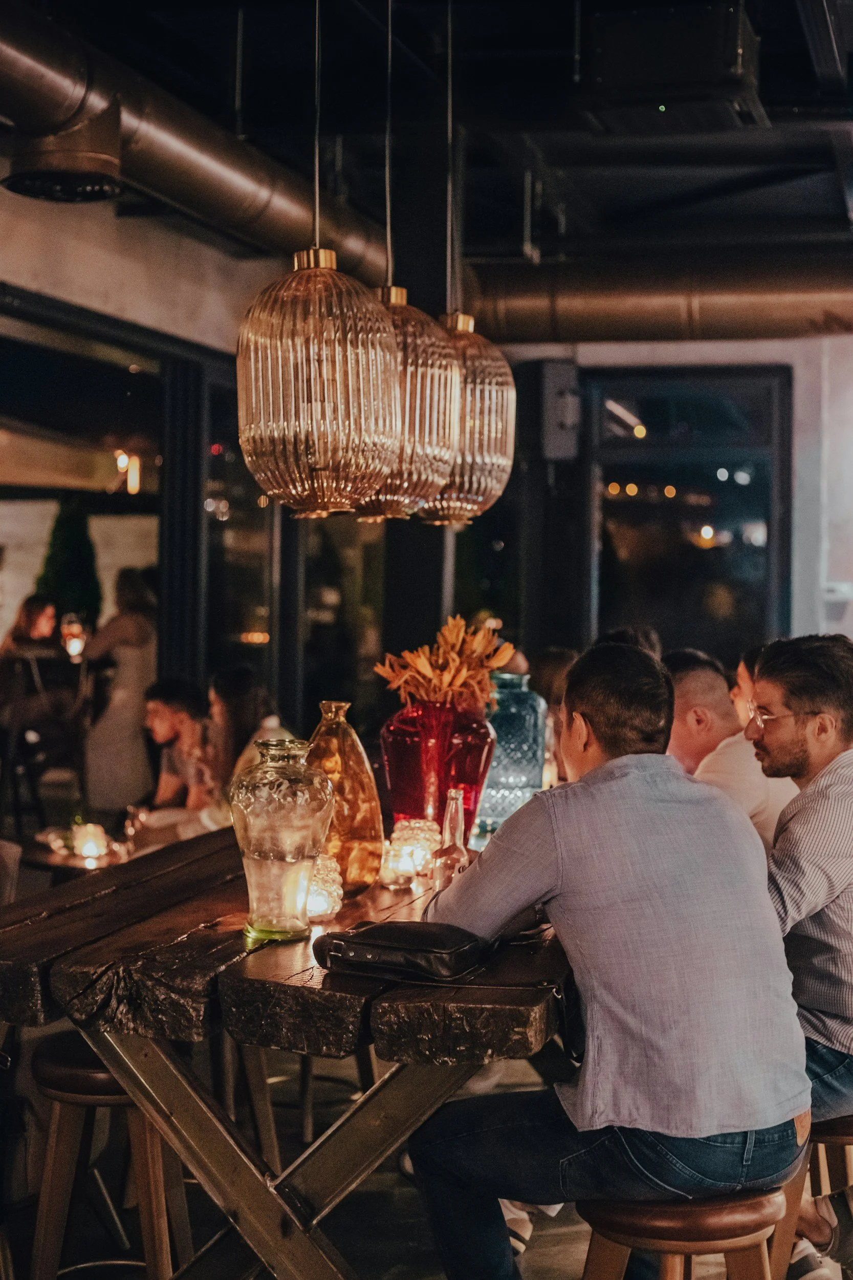 People sitting at a bar in a dimly lit restaurant or bar, with hanging pendant lights, decorative vases, and glasses on the counter. Large windows are visible in the background.