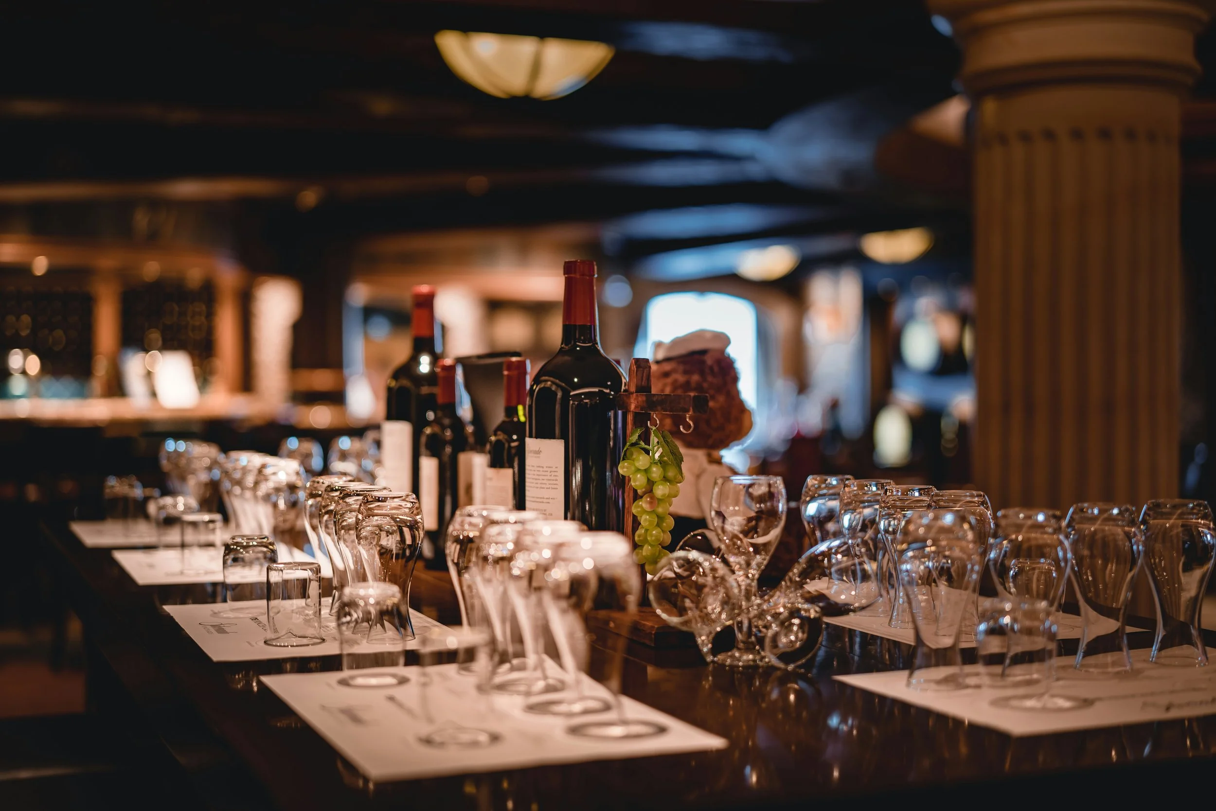 A restaurant table set with empty wine glasses, bottles of wine, green grapes, and menus with a dimly lit wood-paneled interior.