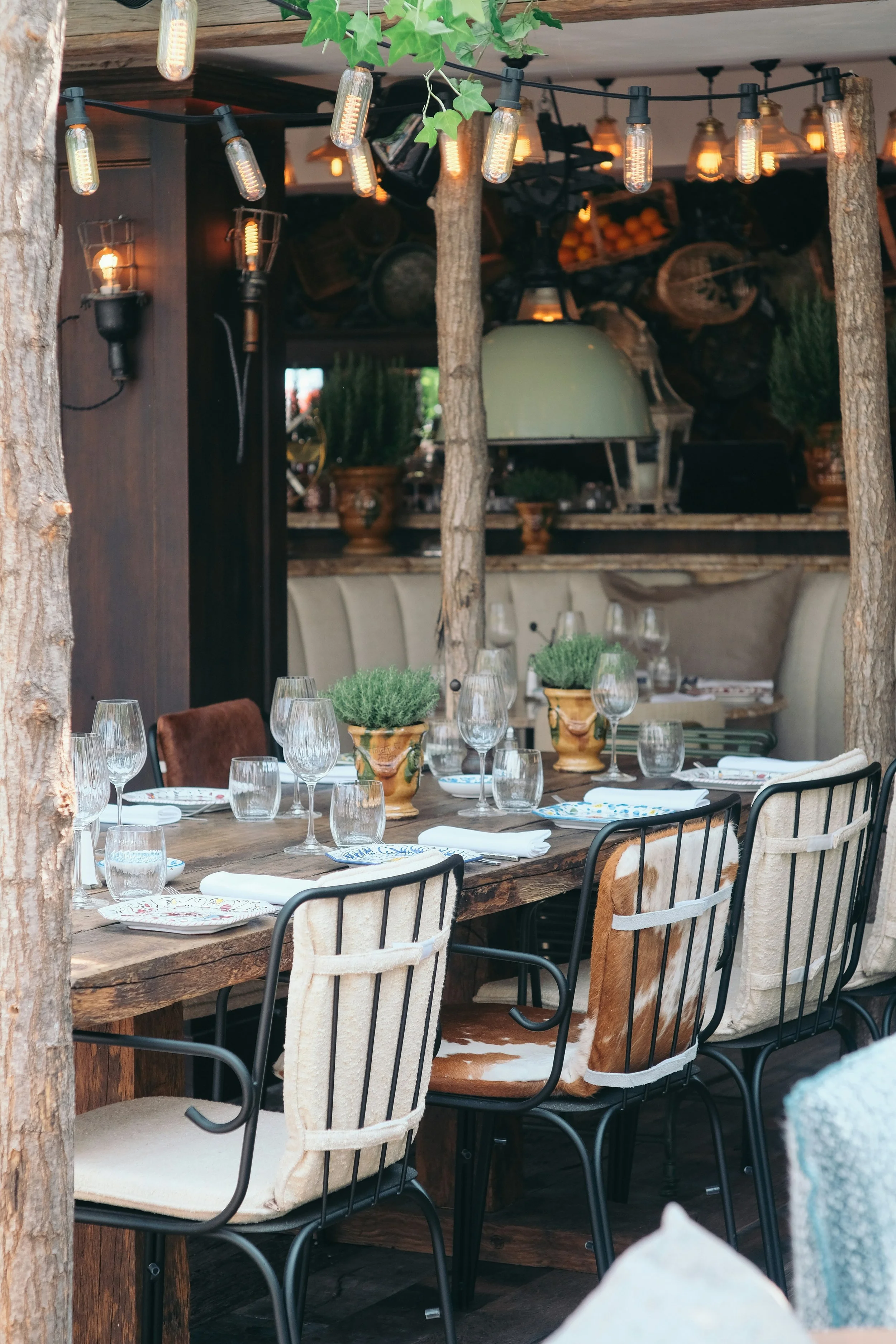 A rustic outdoor dining area with a large wooden table set with wine glasses, plates, and vases with greenery. The background shows a cozy interior with warm lighting, decorative pots, and a pizza oven. String lights hang overhead, and tree trunks are part of the décor.