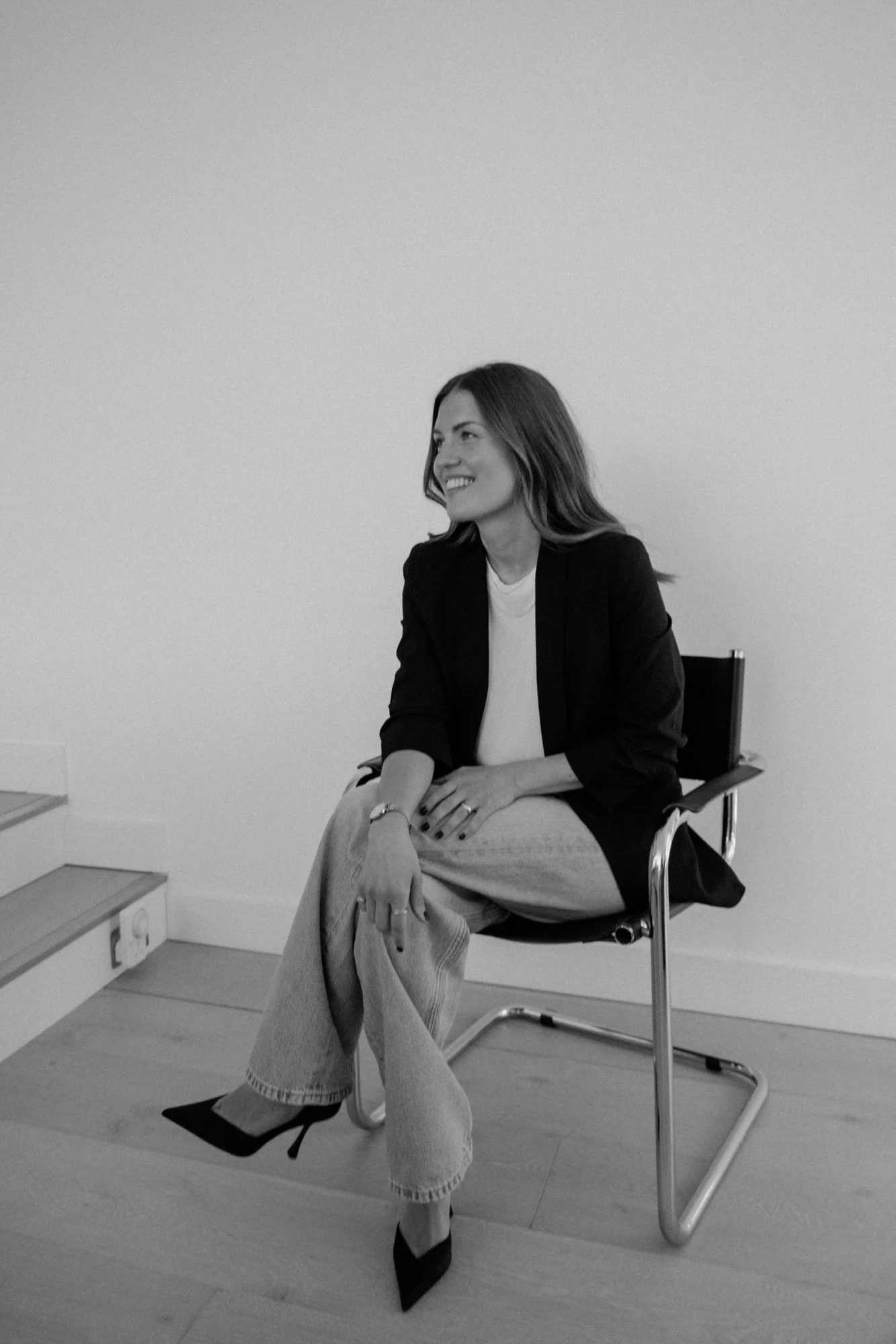 A woman sitting on a black office chair with a black blazer and high heels, smiling and looking to her left, in a room with plain white walls, a wooden floor, and a staircase.