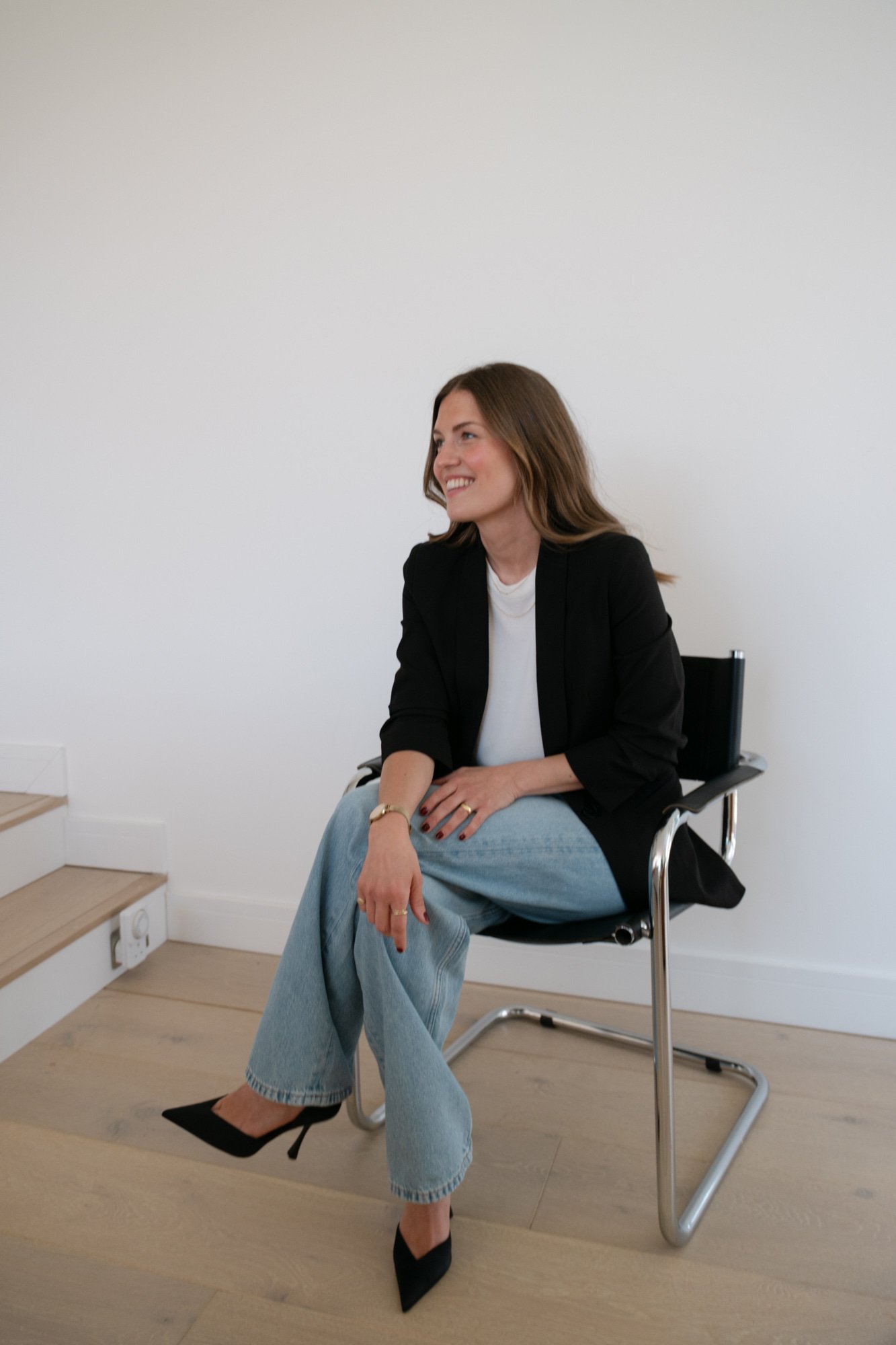 A woman with shoulder-length brown hair, smiling, is sitting in a black office chair against a plain white wall. She is wearing a black blazer, white top, light blue jeans, and black high heels. She has jewelry on her fingers and wrist, with her left hand resting on her knee and her right hand on her lap. There are stairs and a power outlet visible next to her.
