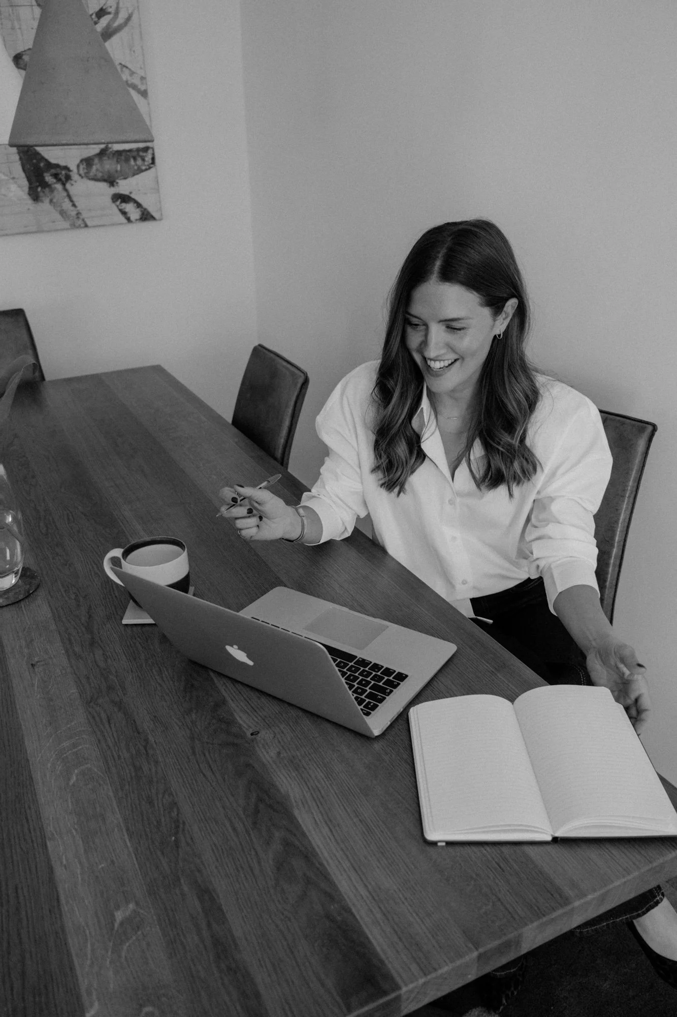 A woman with long hair and a white shirt sitting at a wooden table with a laptop, open notebook, mug, and smartphone, smiling and looking down, in a room with a wall art piece of a lobster in the background.