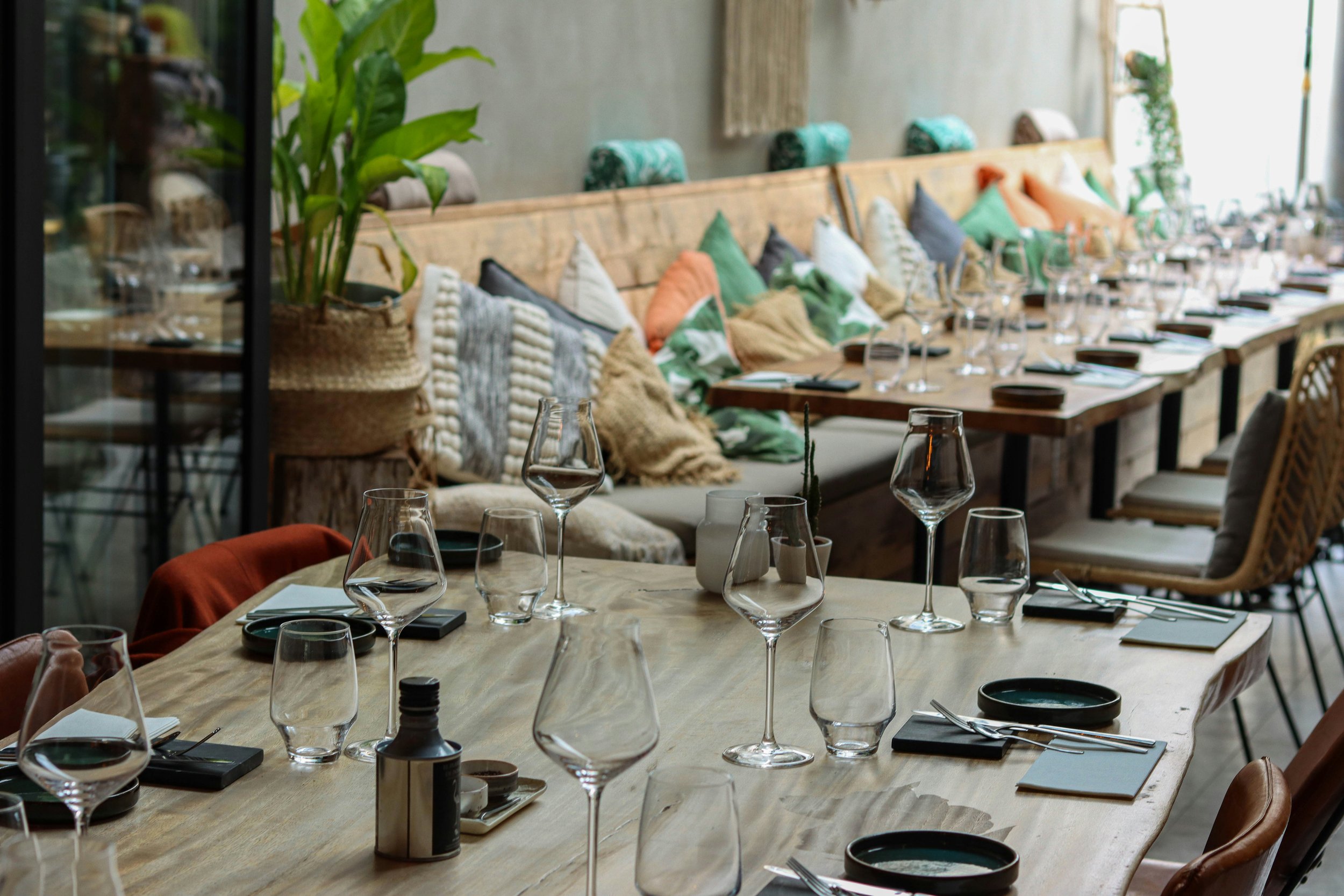 A restaurant dining area with a long wood table set with wine glasses, small sauce dishes, and napkins. The background features a wall with decorative pillows on a bench and some indoor plants.