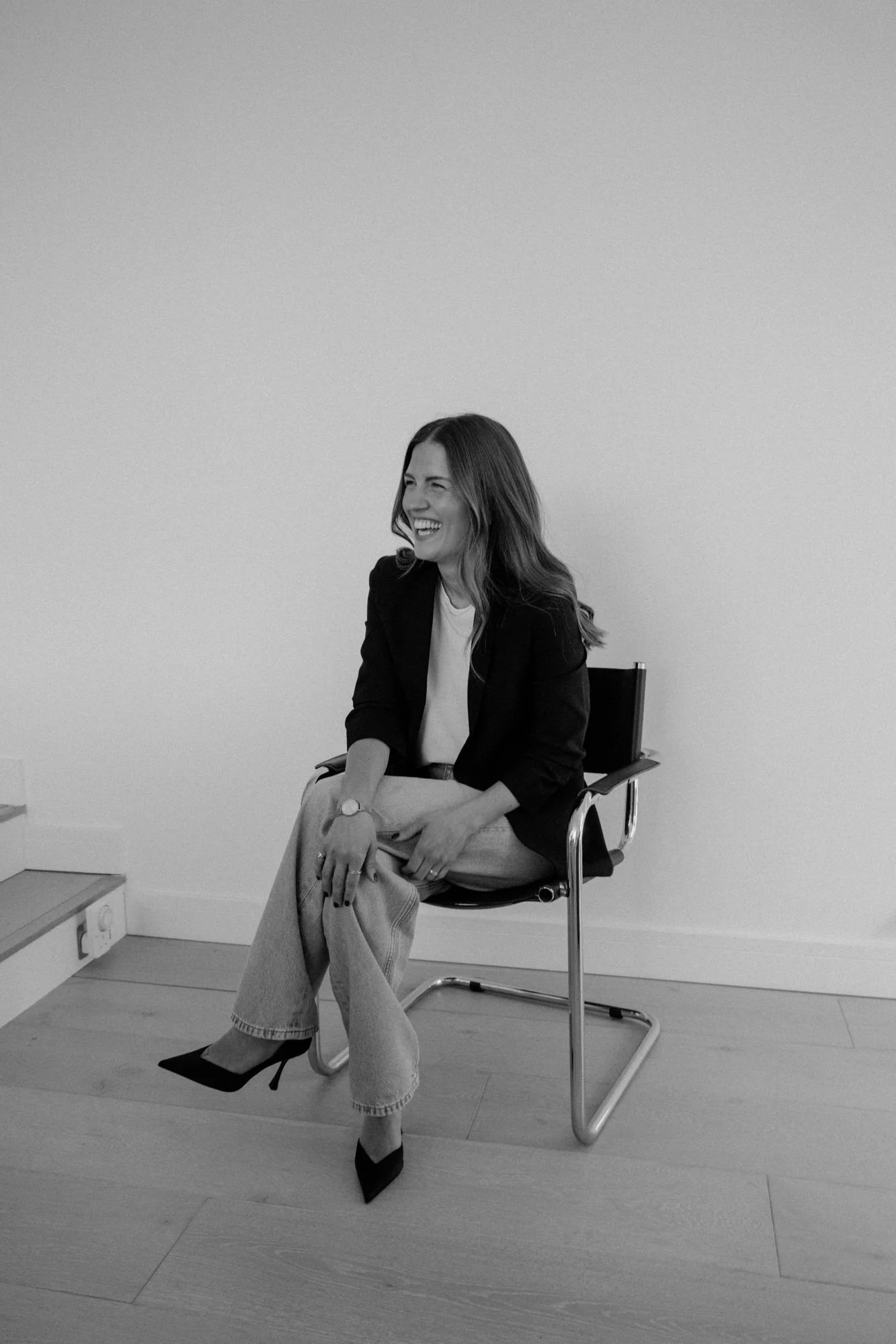 A woman with long hair, wearing a blazer and light-colored pants, sitting on a black office chair, smiling and laughing, in a minimalistic room with plain wall and wooden floor.