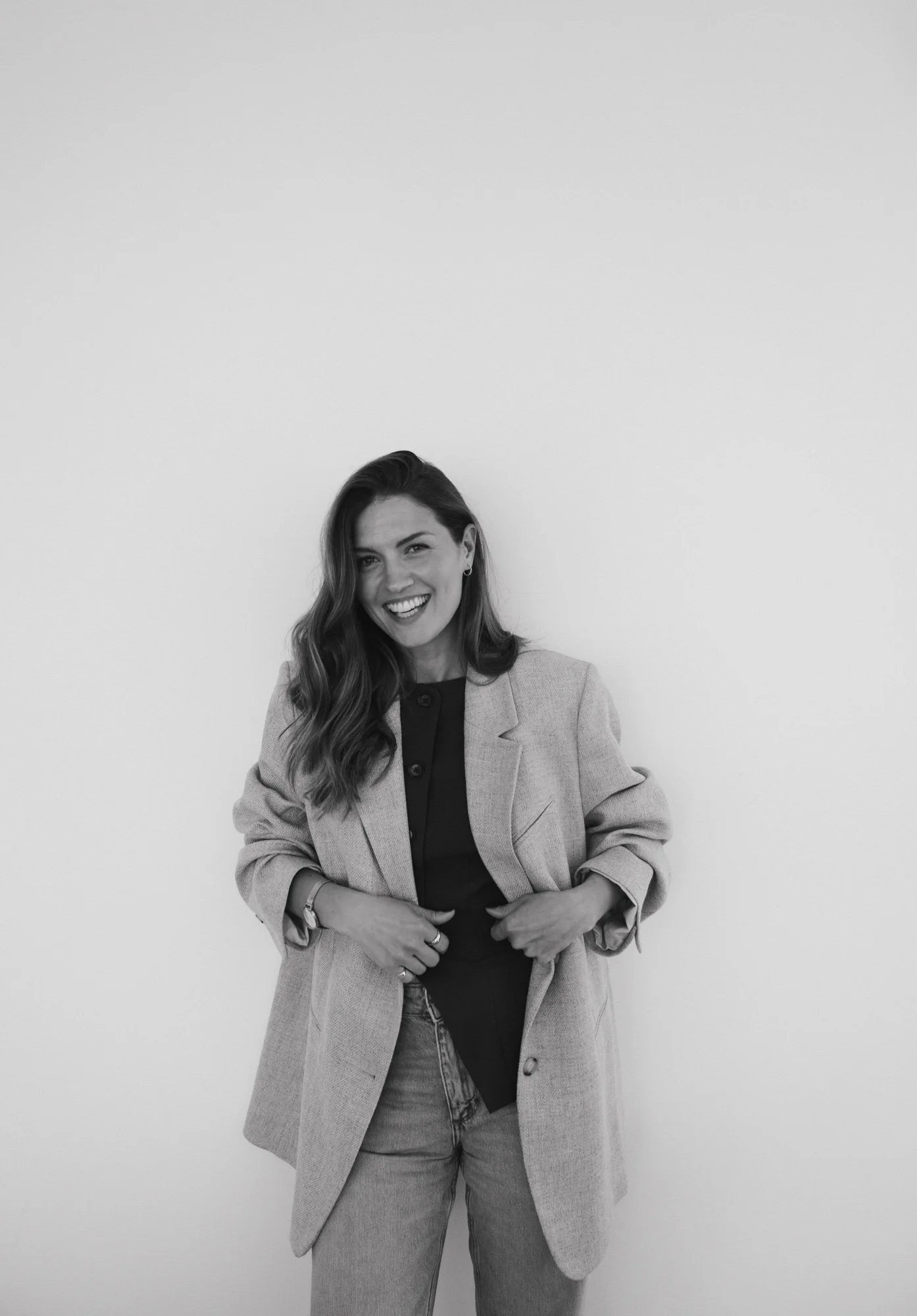 A woman with long wavy hair smiling and looking at the camera, wearing a light-colored blazer over a dark shirt, with her hands adjusting her blazer, standing against a plain white background.
