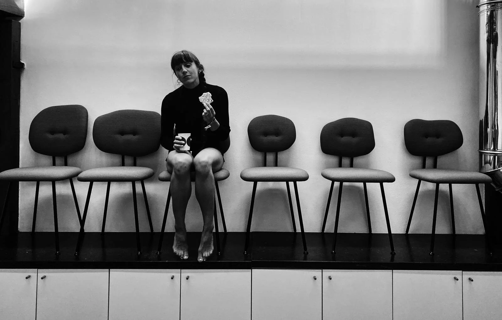 A woman sitting alone on a row of chairs in an empty room, holding a cup and a cookie. The room has a plain wall and a polished metal pipe on the right. The woman is barefoot and looks contemplative.