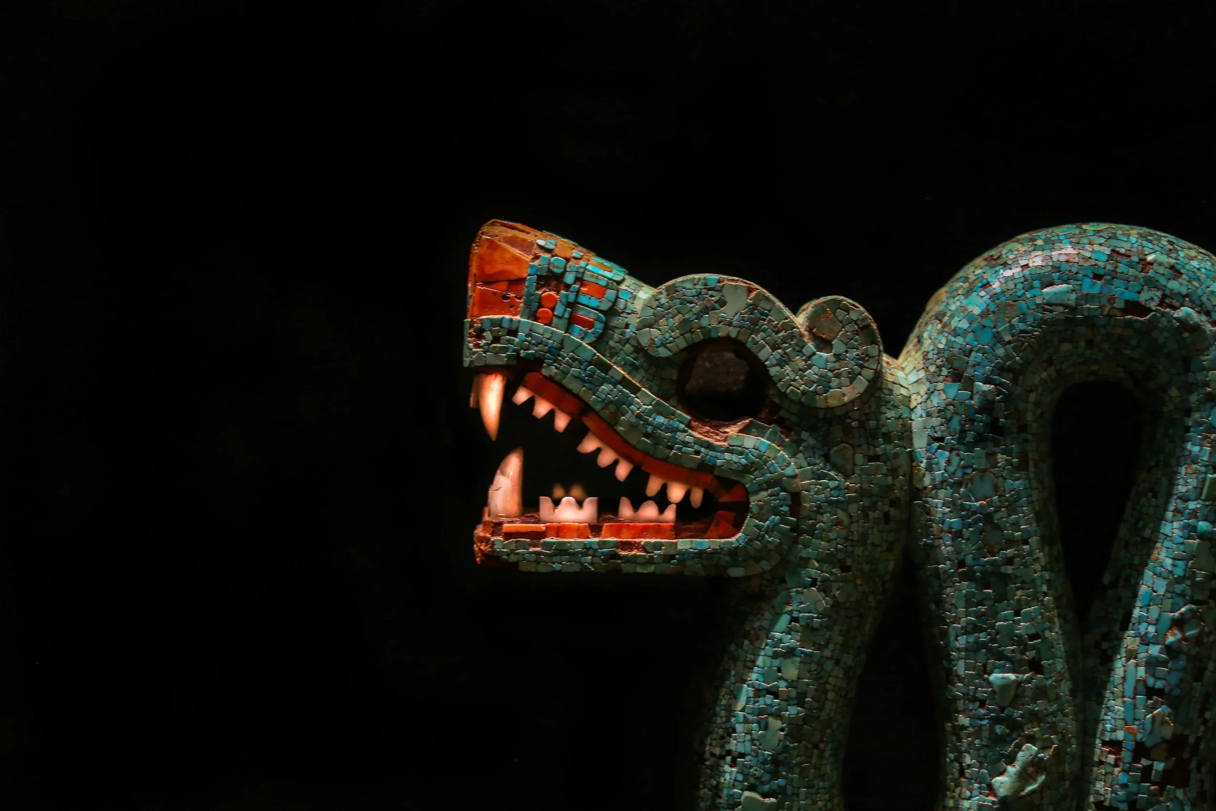 A mosaic sculpture of a dragon's head, showing sharp teeth and an open mouth against a black background.