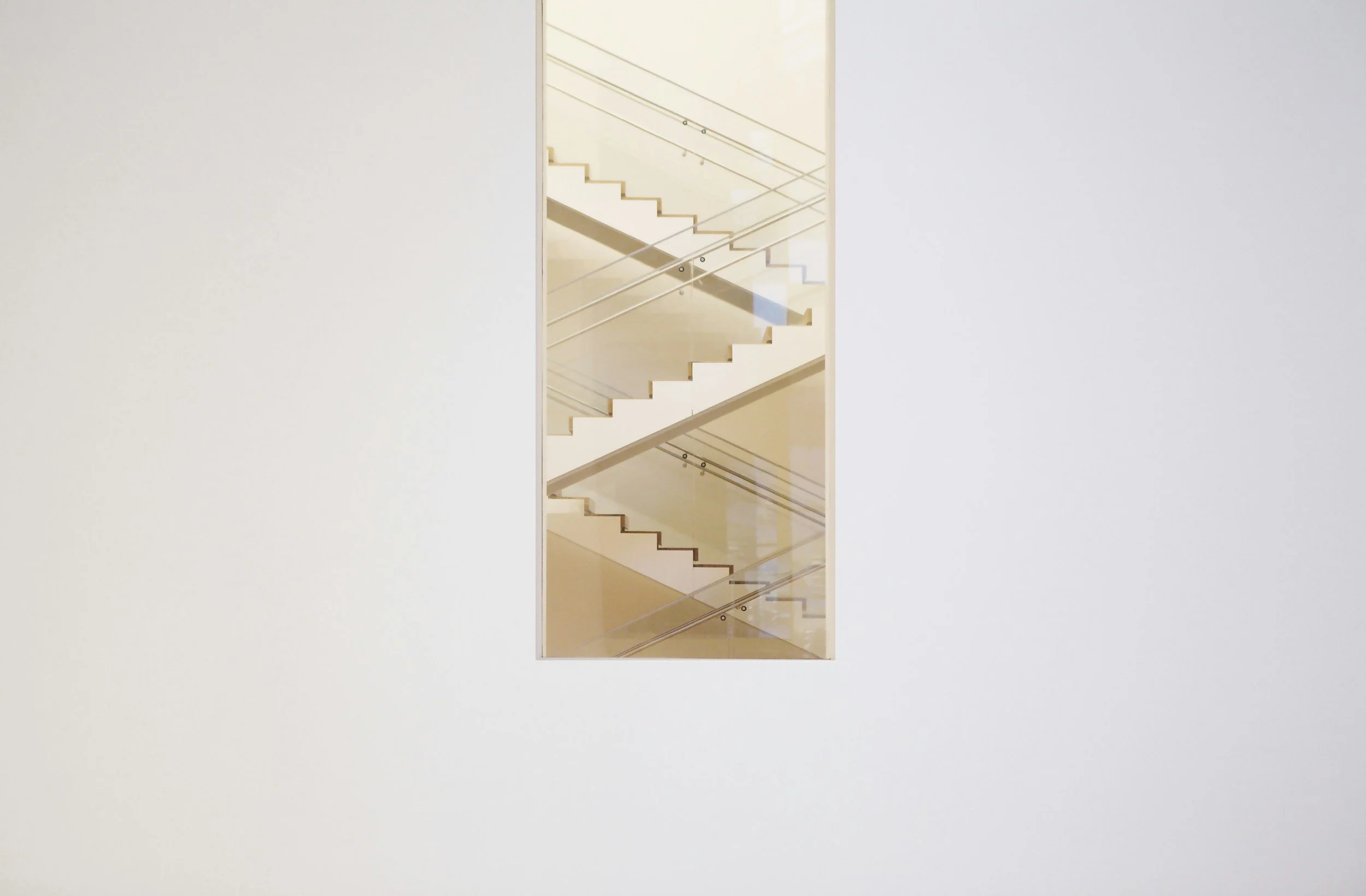 A modern staircase with white steps and glass railings, viewed through a narrow rectangular window in a white wall.
