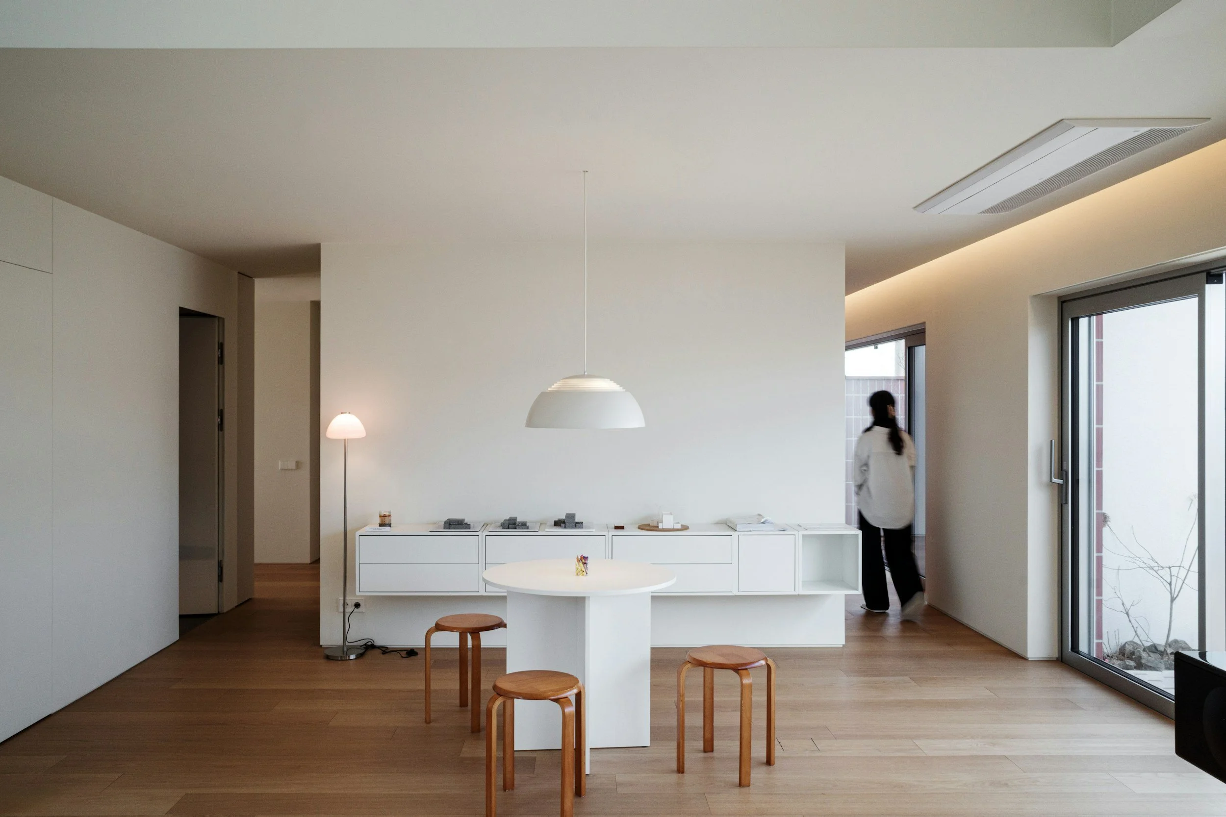 Interior of a modern, minimalist room with white walls, a white dining table, wooden stools, and a white sideboard, with a person walking near the side door and natural light coming in through large windows and a glass sliding door.
