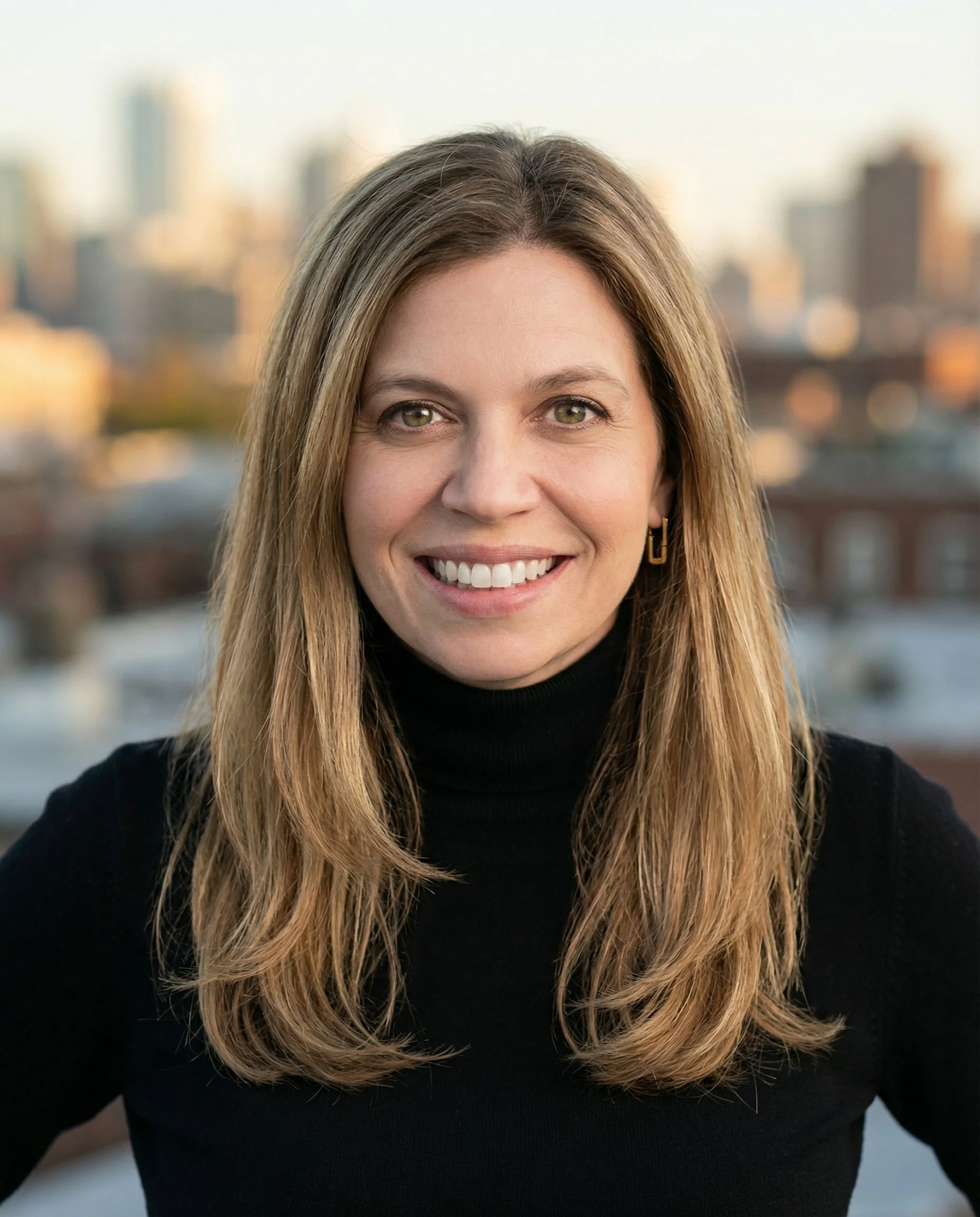 Close-up of a woman smiling, with long wavy blonde hair, wearing a black turtleneck and gold earrings, with a cityscape blurred in the background.