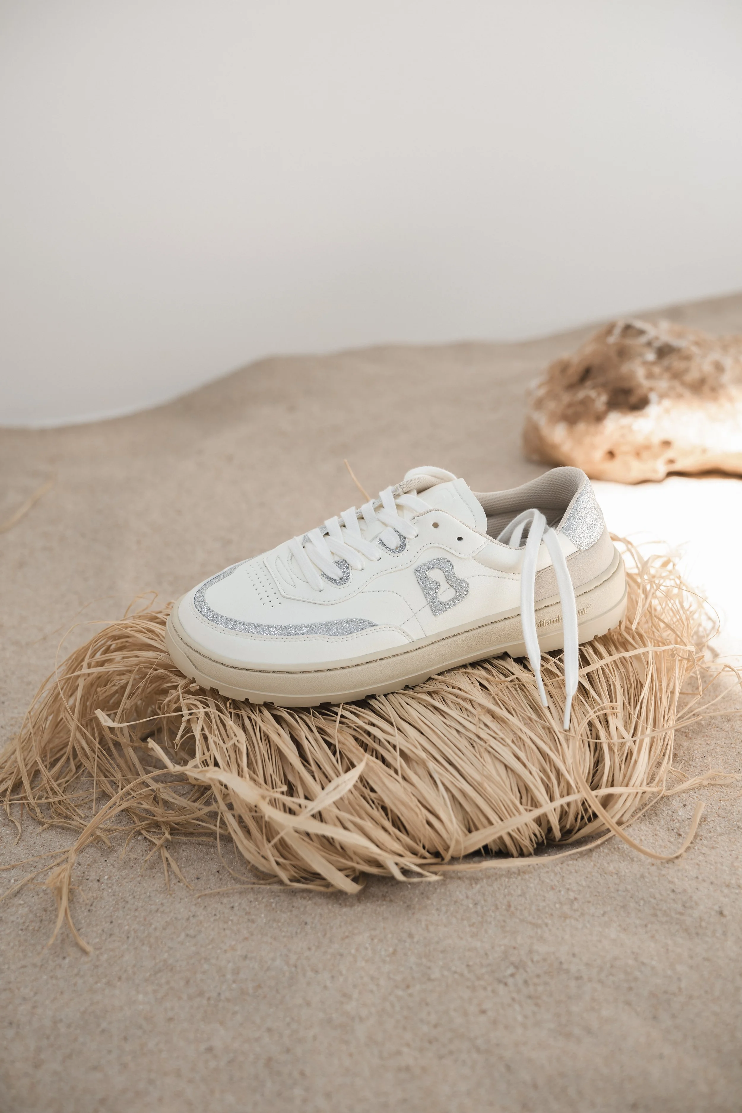 Zapatillas blancas con detalles plateados en un saco de yute sobre arena clara junto a una roca.