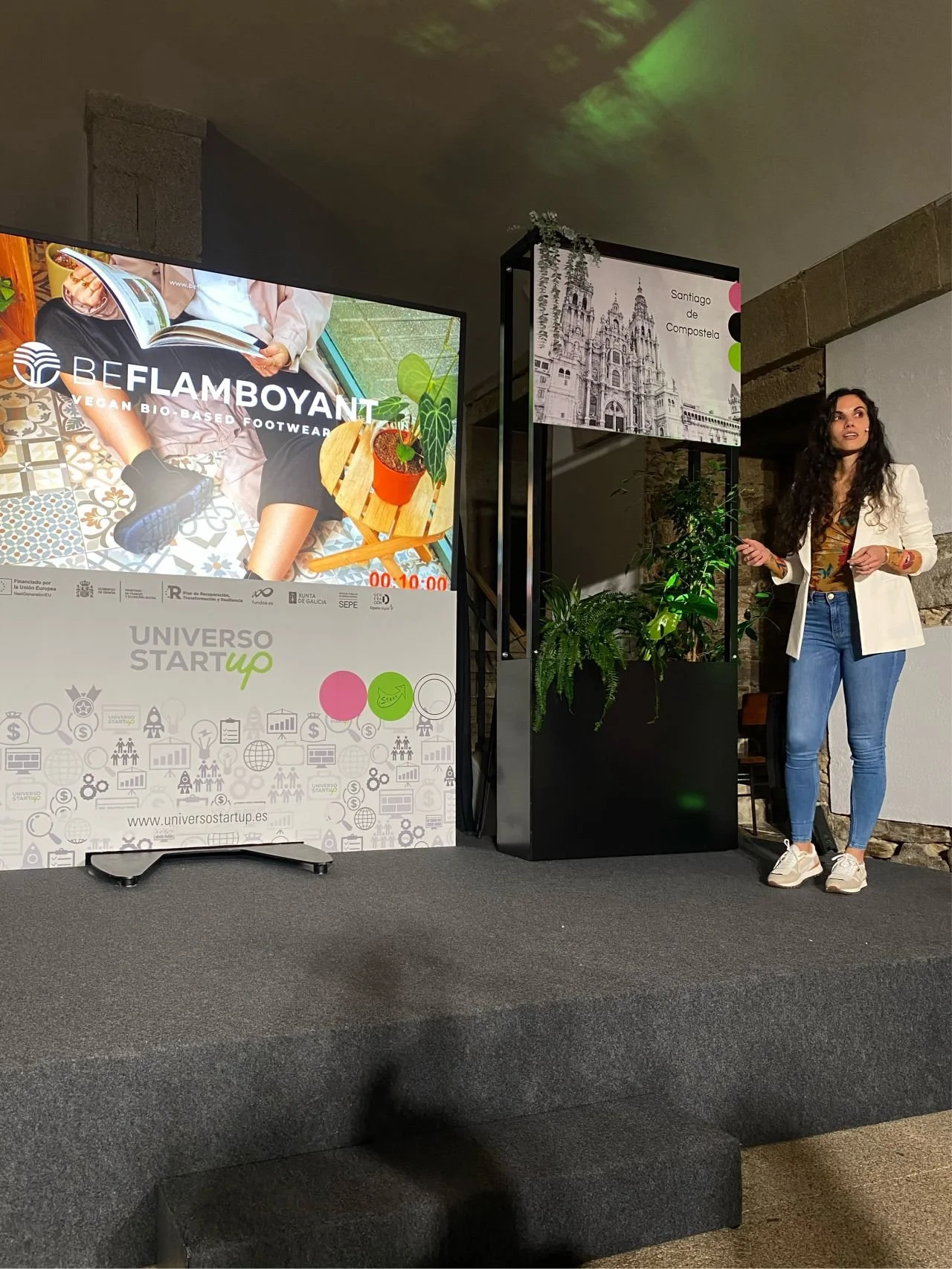 Presentación en una conferencia sobre emprendimiento y sostenibilidad, con una mujer de cabello rizado y oscuro con vestimenta casual y fondo de plantas.