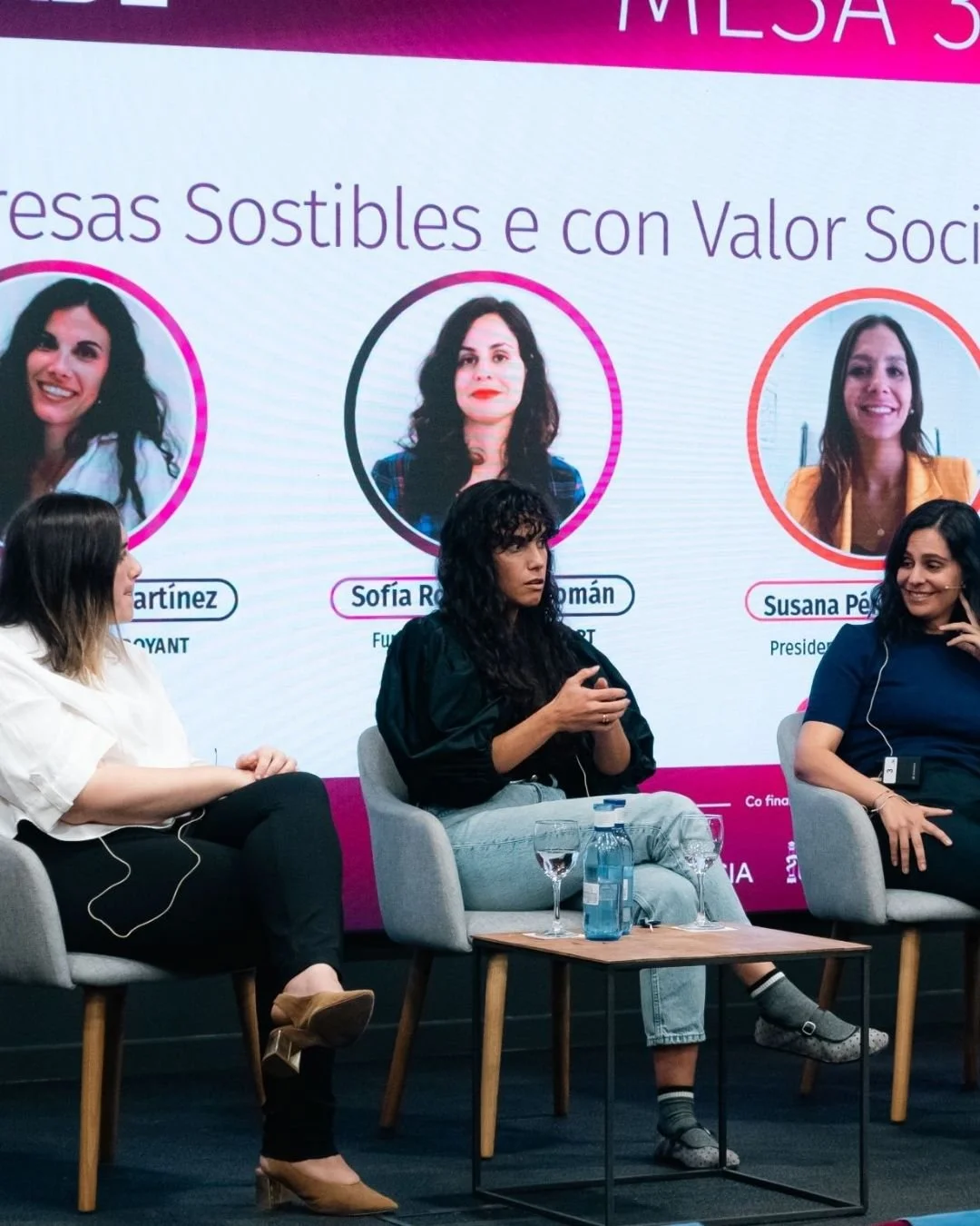 Tres mujeres participan en un panel de discusión en una conferencia, sentadas en sillones frente a un cartel grande que muestra retratos y nombres de otras mujeres, con fondo rosado y blanco.