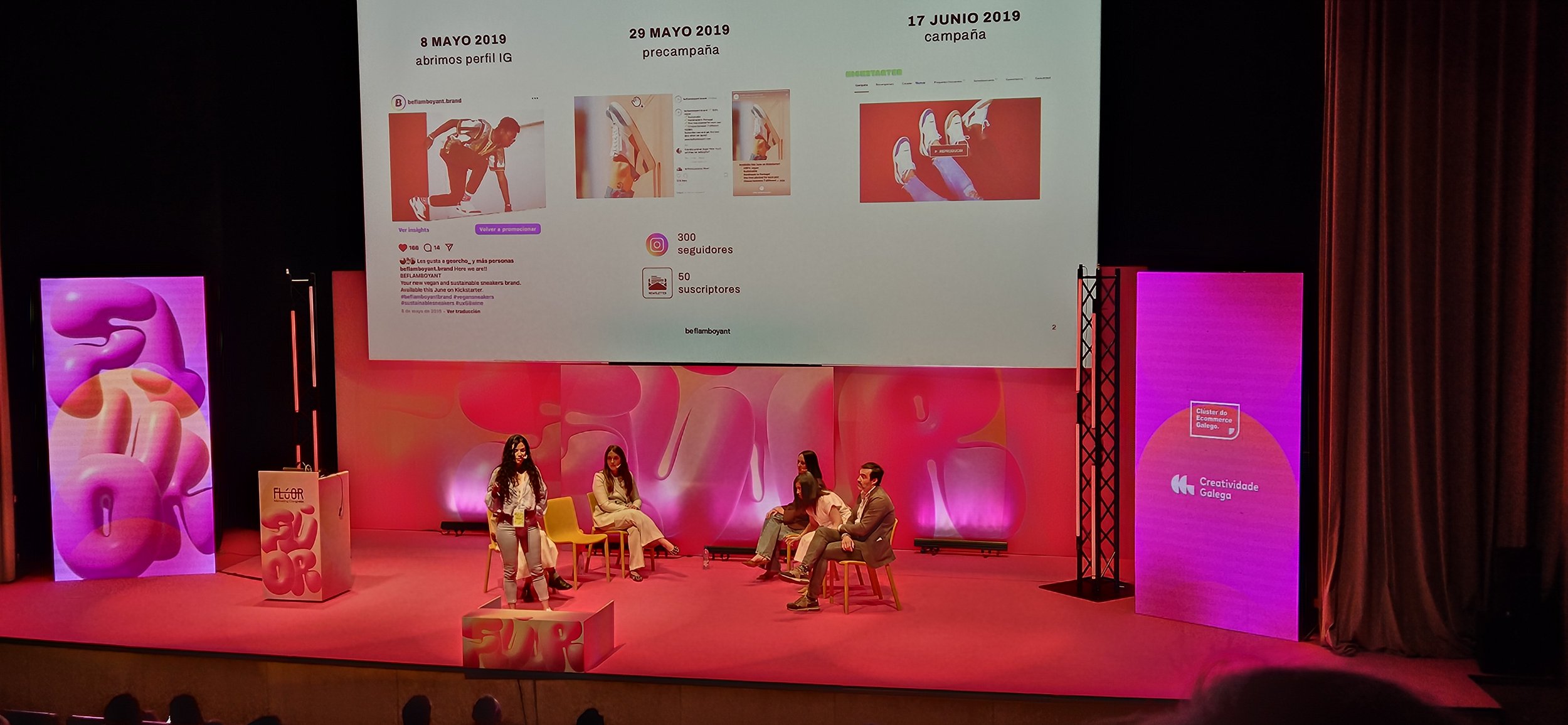 Escenario con cinco personas sentadas y una de pie, en medio de una conferencia o panel en un evento. Detrás, pantalla grande mostrando información con fechas y ejemplos de publicaciones en redes sociales. A la izquierda y derecha, pantallas verticales con diseños coloridos y un cartel con el texto 'Creatividade Galega'.