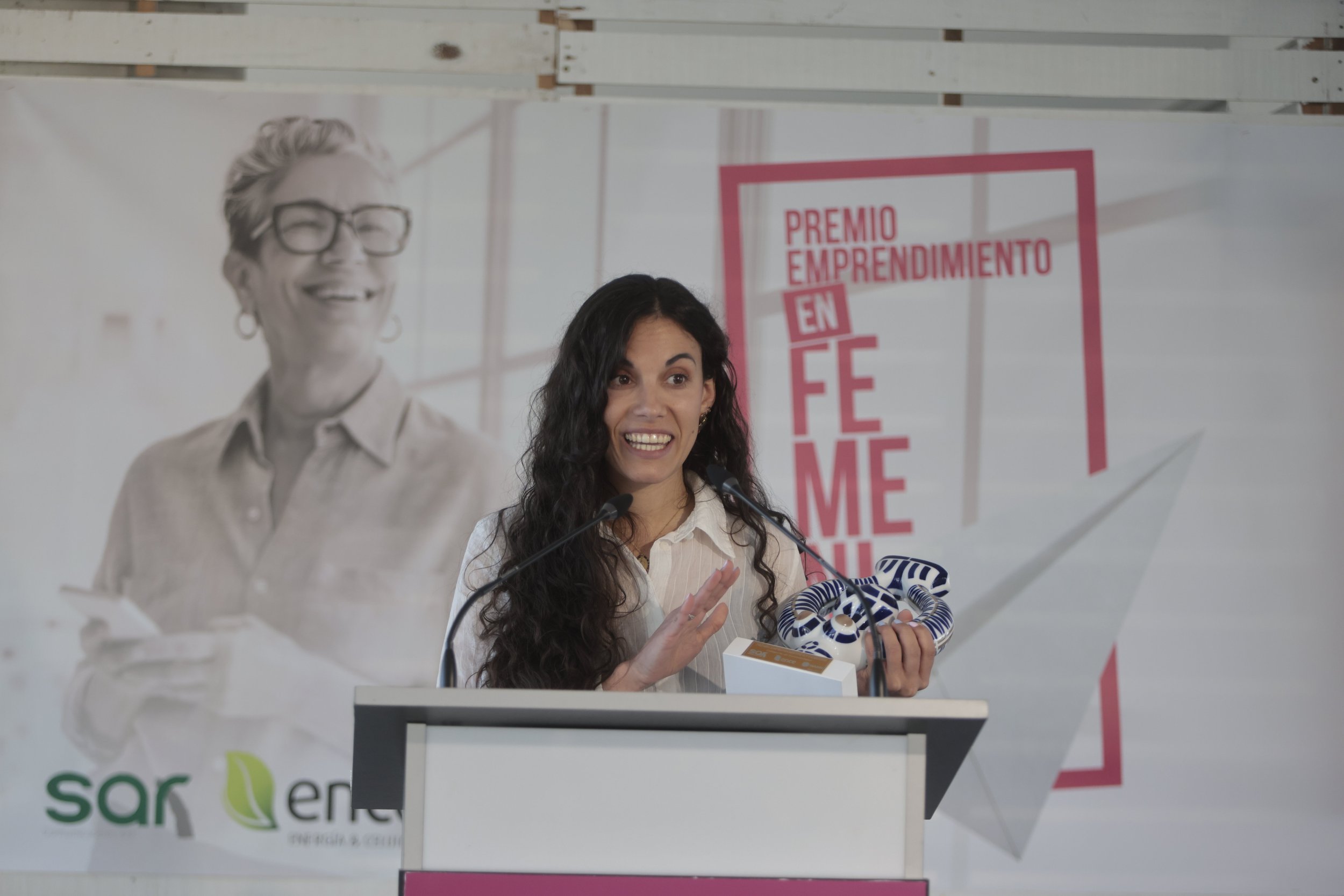 Una mujer con cabello rizado oscuro y vestida con blusa blanca está dando un discurso o mensaje en un podio durante un evento. En el fondo se ve un cartel con letras en color rosa y rojo que dice 'Premio Emprendimiento en F' y una imagen grande de una mujer mayor con gafas y cabello gris. Hay micrófonos sobre el podio y una mano sosteniendo un objeto con rayas azules y blancas.