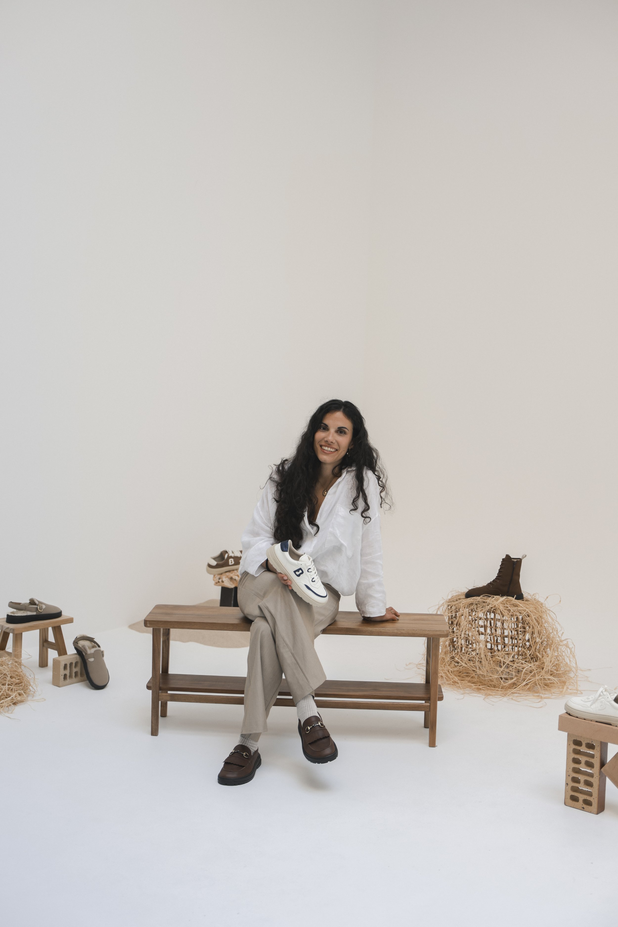 Mujer sonriendo, sentada en banco de madera, rodeada de calzado y decoraciones de estilo rústico en un ambiente minimalista con paredes blancas.
