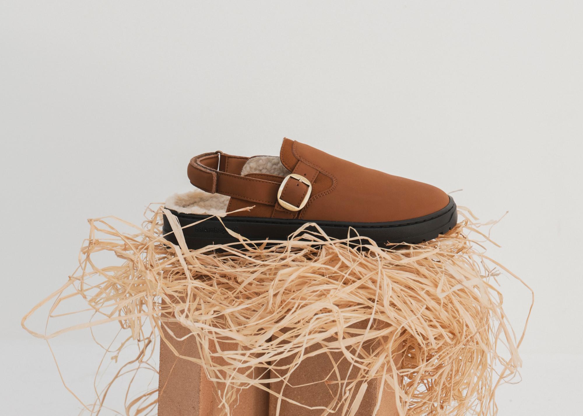 Zapato de piel marrón con forro de felpa, colocado sobre una estructura de madera y papel decorativo enmarañado.