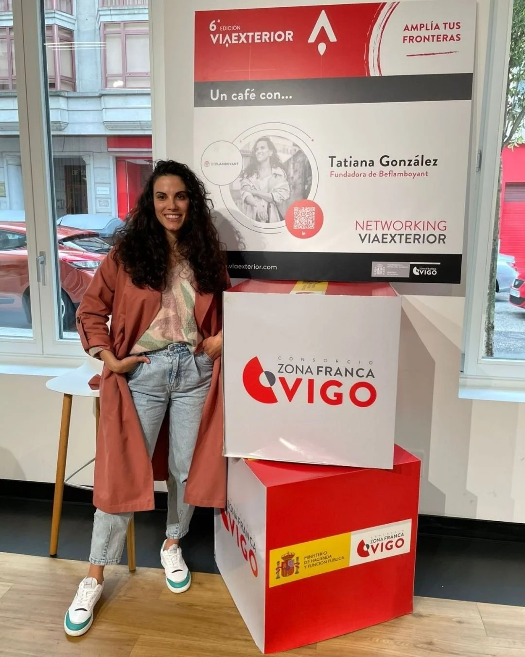 Una mujer sonriente con cabello rizado, vestida con un abrigo color terracota, camiseta clara y jeans, posa junto a cajas con logotipos de Zona Franca Vigo y del Ministerio de Hacienda en un espacio interior con ventanas y vehículos afuera.