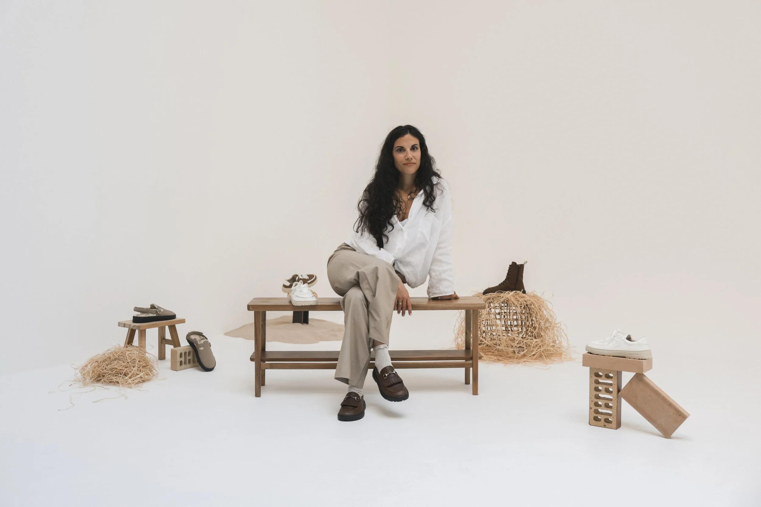 Mujer con ropa blanca y pantalones beige, sentada en un banco de madera rodeada de calzado y objetos decorativos de madera y cuerda, en un fondo blanco.