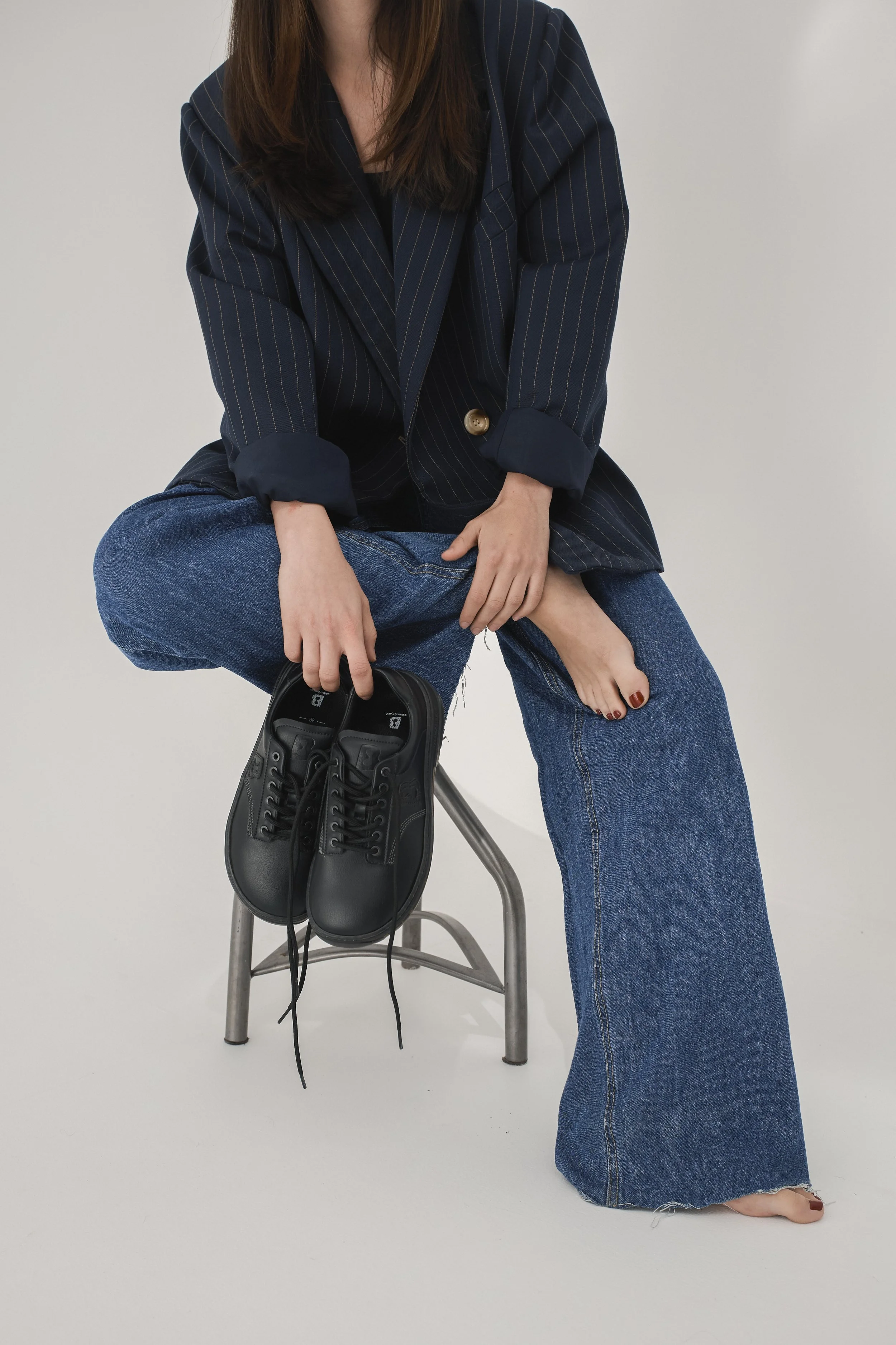 Persona de pie con una pierna cruzada, sosteniendo un zapato negro con cordones, con una silla de metal en el fondo, viste pantalones de mezclilla anchos y una chaqueta azul con rayas, sin mostrar el rostro.