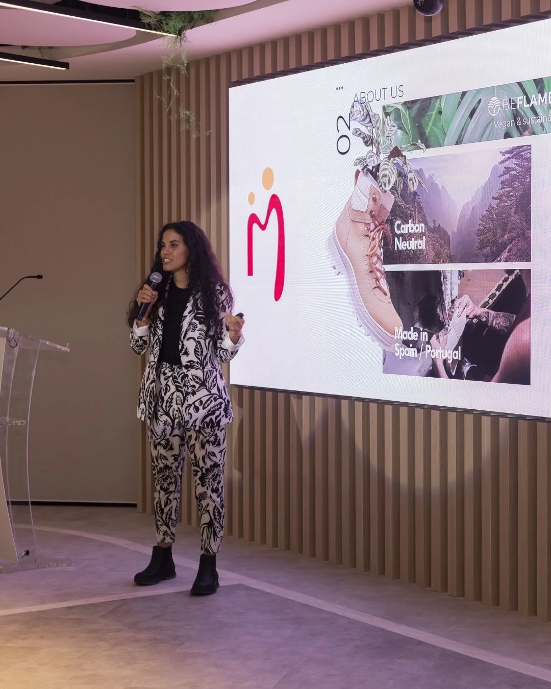 Una mujer con vestido de pantalón y chaqueta con estampado floral negro y blanco da una presentación frente a una pantalla grande que muestra información sobre una marca de zapatos veganos y sostenibles, con detalles sobre sus características y origen.