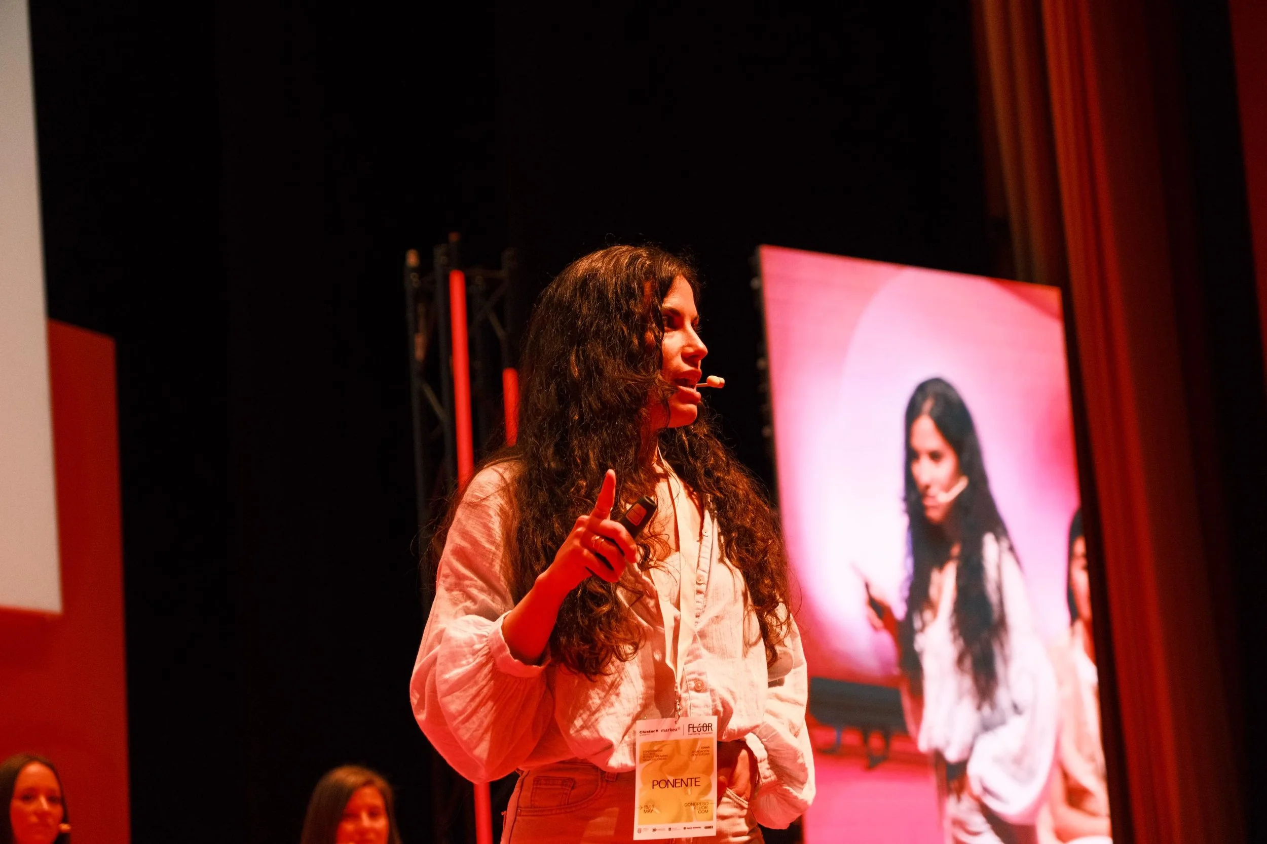 Mujer con cabello rizado oscuro y camisa clara hablando con micrófono en frente de una pantalla grande en un escenario. Personas sentadas en el público