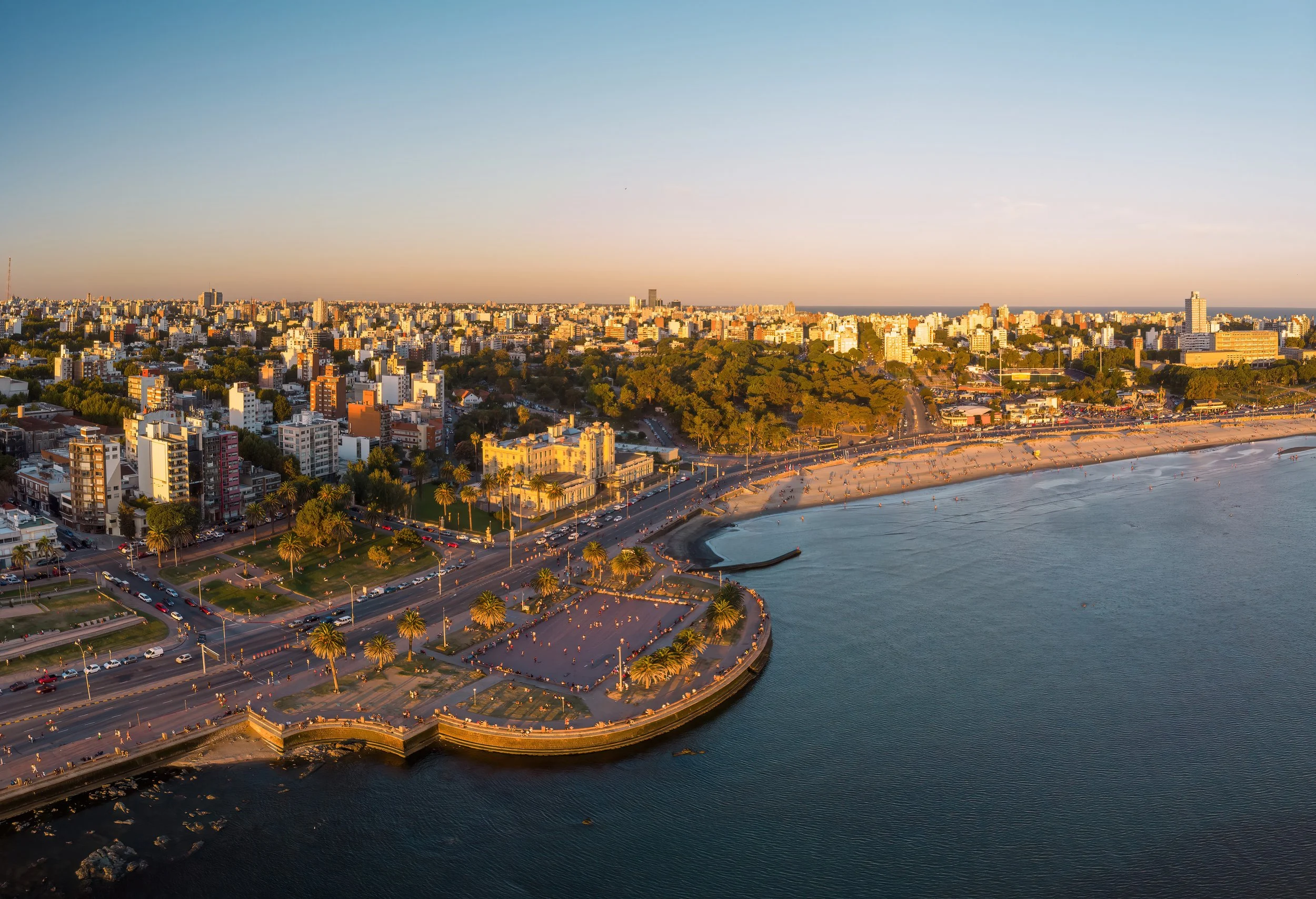A panoramic aerial view of a city coastline at sunset, featuring a beach with people, a promenade lined with palm trees, and numerous buildings and skyscrapers in the background.