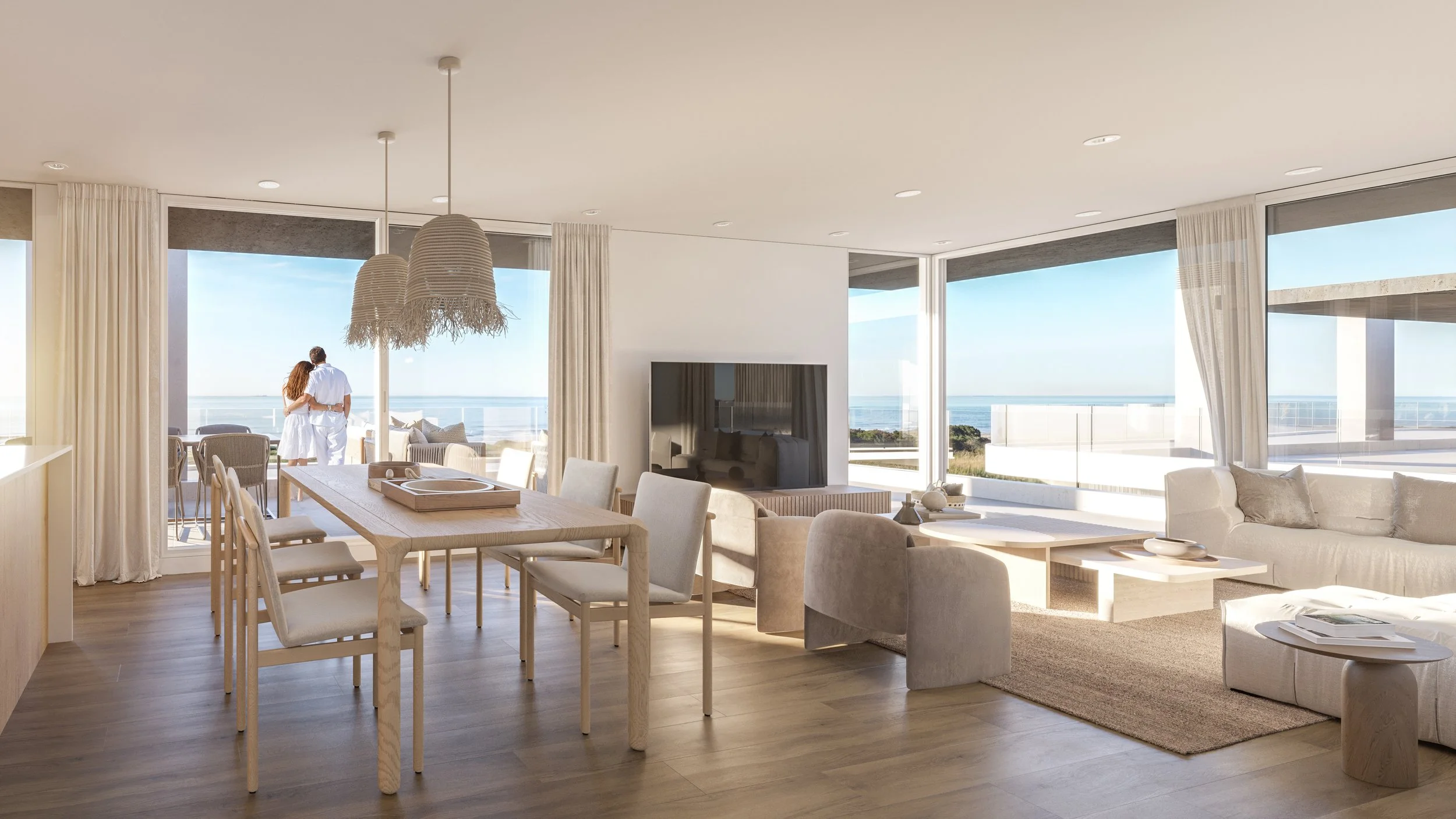 Bright, airy living room and dining area with large windows revealing an ocean view, modern white and beige furniture, a flat-screen TV, and a couple standing on the balcony.