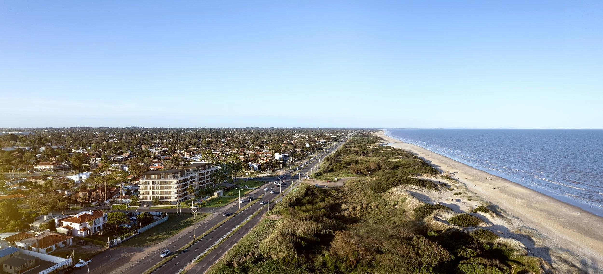 Aerial view of a coastal city showing residential buildings, a road, and a sandy beach alongside the ocean on a clear day.