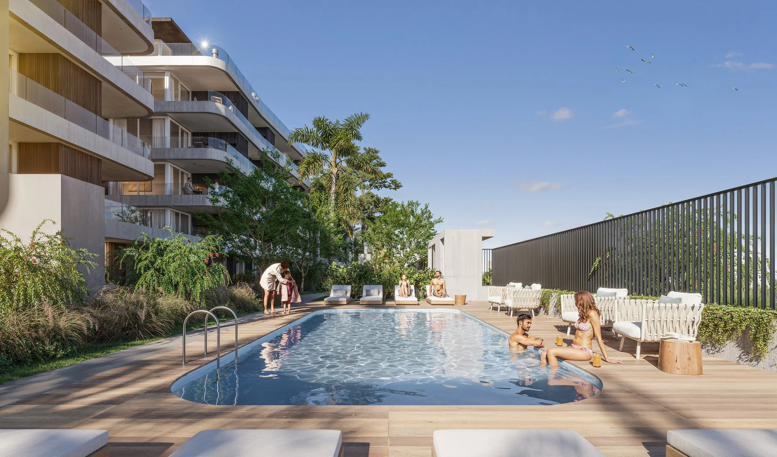 People enjoying a swimming pool area outdoors with lounge chairs, surrounding greenery, and modern apartment buildings in the background under a clear blue sky.