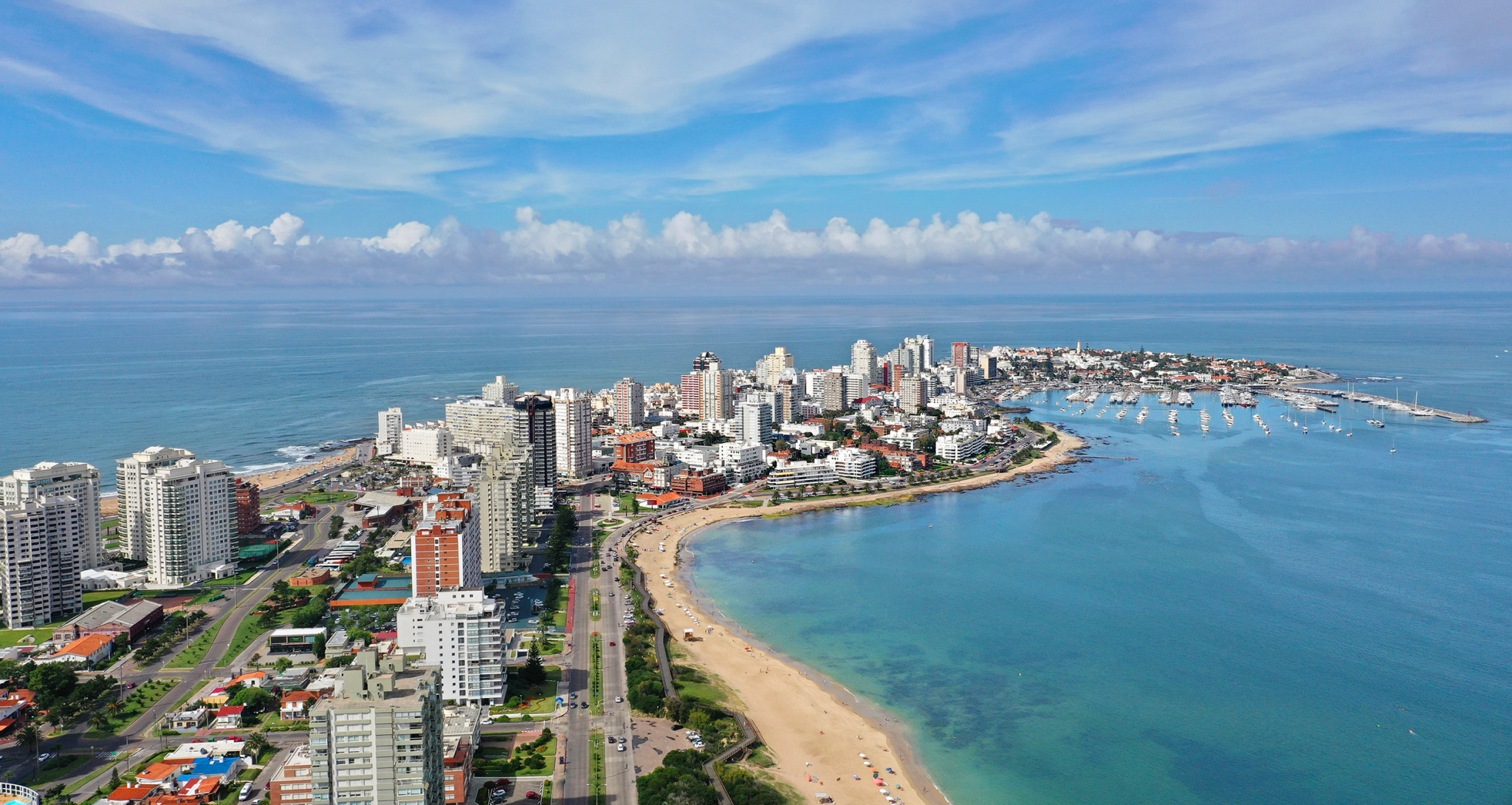 A coastal city skyline with high-rise buildings along the waterfront, a sandy beach, and a marina with boats, under a blue sky with scattered clouds.