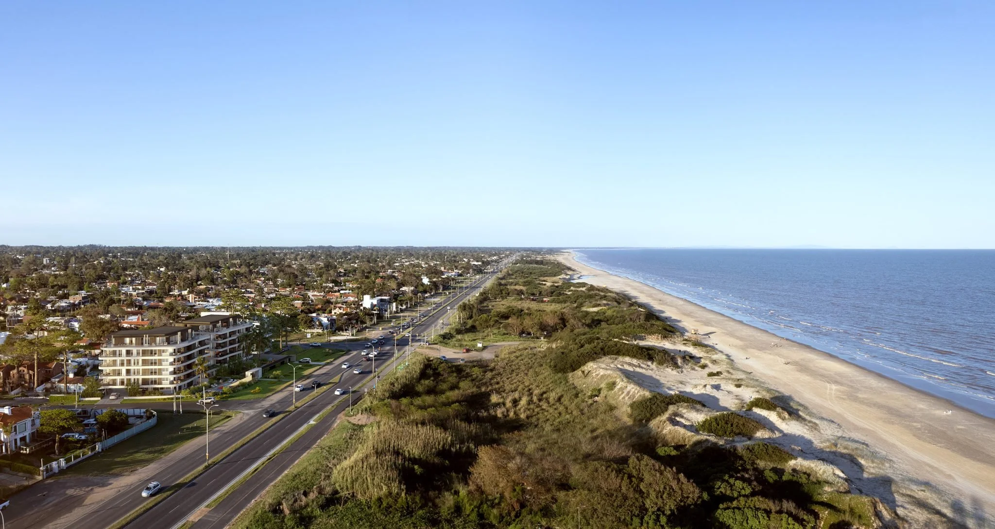 A coastal city view with houses, a road, and a beach along the shoreline, with the ocean on the right and a clear blue sky above.