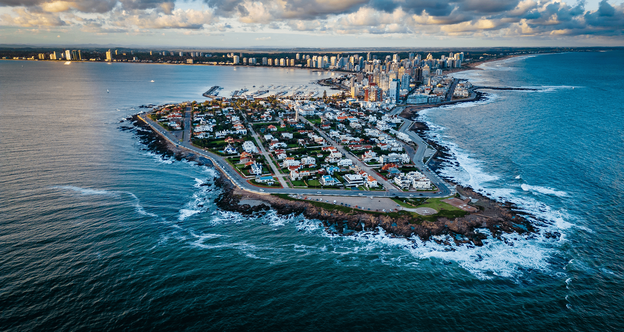 Aerial view of a coastal city with a peninsula featuring residential homes, a helipad, and high-rise buildings along the shoreline, with water and clouds visible in the background.