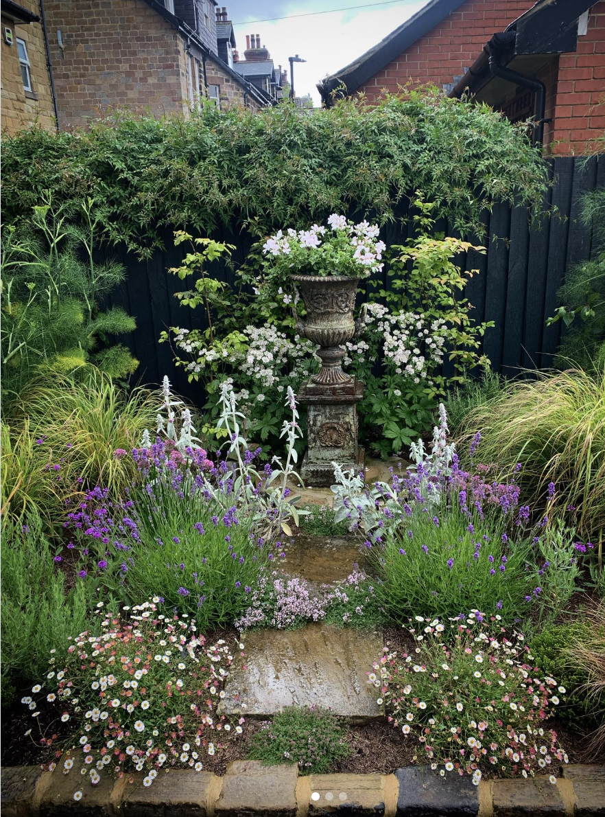 A colorful garden with various blooming flowers, a stone pathway, and an ornate stone planter in the center, surrounded by lush green foliage and plants.