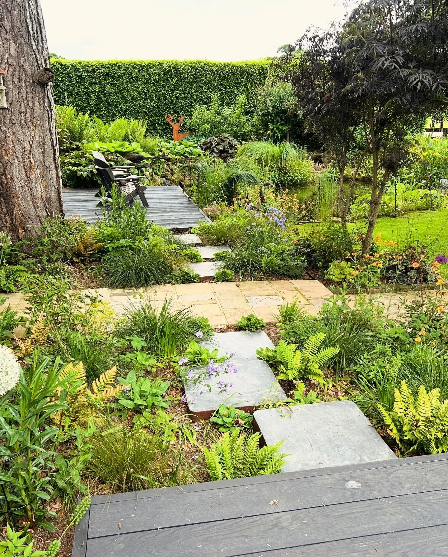 A lush garden scene with a wooden deck and pathway, surrounded by various green plants, trees, and colorful flowers. There is a large tree trunk on the left, a pond on the right, and a small decorative deer statue in the background. The garden appears well-maintained and peaceful.