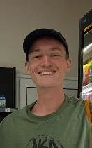 A young man smiling in a retail store, wearing a baseball cap and green shirt.