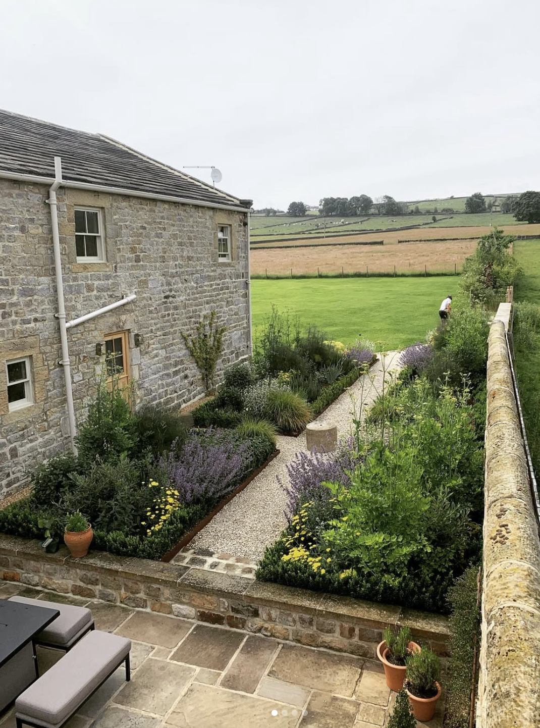 View of a stone house garden with a gravel path, shrubs, and flowers, overlooking a green lawn and open fields, with a person tending to plants in the background.