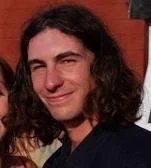 A person with long, curly dark hair smiling outdoors.