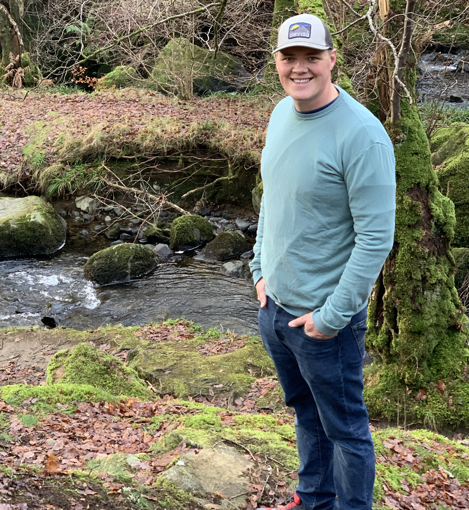 A young man in a teal long-sleeve shirt, jeans, and a gray cap, smiling and standing outdoors near a stream with moss-covered rocks and trees.