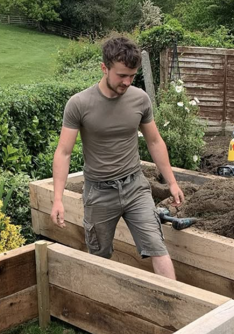 A young man in a gray t-shirt and shorts working in a garden, standing near a wooden raised bed with a gardening tool on the dirt.