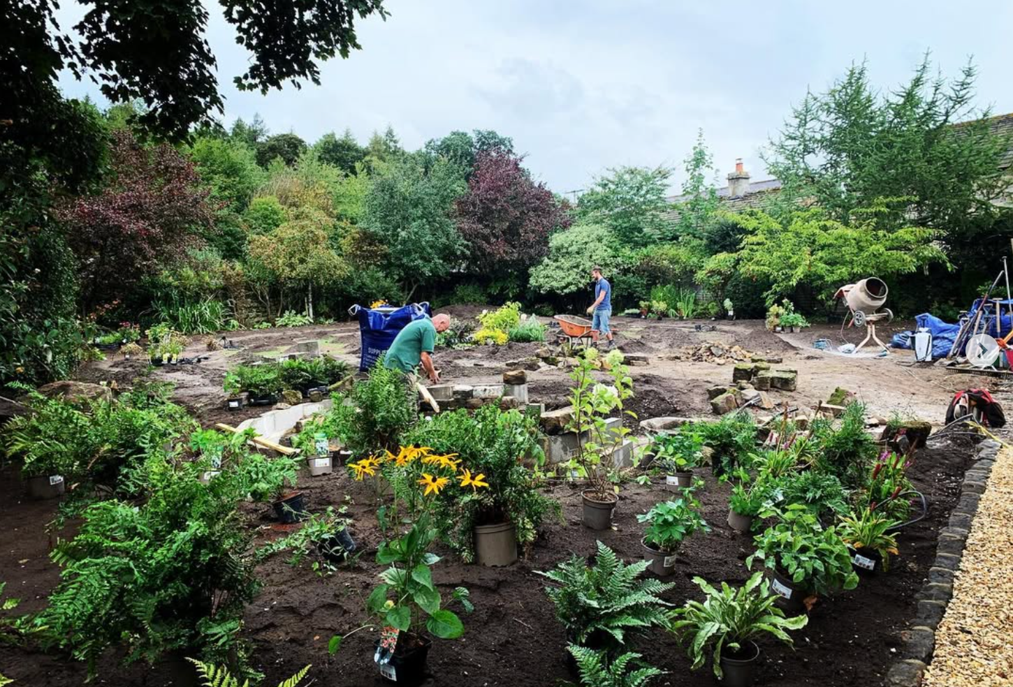 Two people working on landscaping or gardening in a backyard, with trees and bushes in the background, some plants in pots, a cement mixer, a wheelbarrow, and gardening tools.