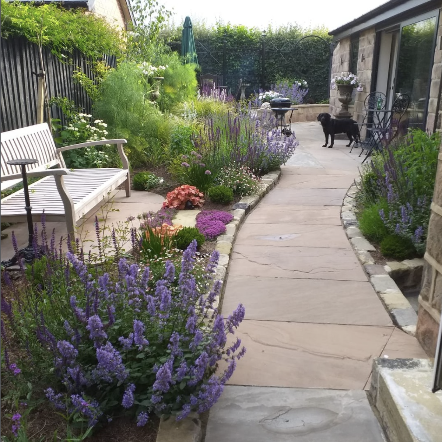 A backyard garden with a stone pathway bordered by colorful flowers and plants. There is a white bench with a small table on the left, and a black dog standing on the right side near outdoor chairs and a grill.